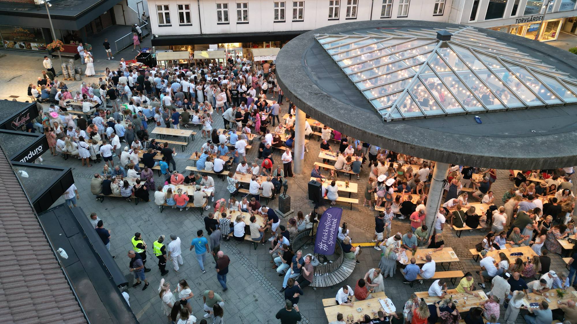 Mange mennesker samlet ved langbord på et torg i Lyngdal sentrum. (Bildebeskrivelsen er laget av en KI-tjeneste)
