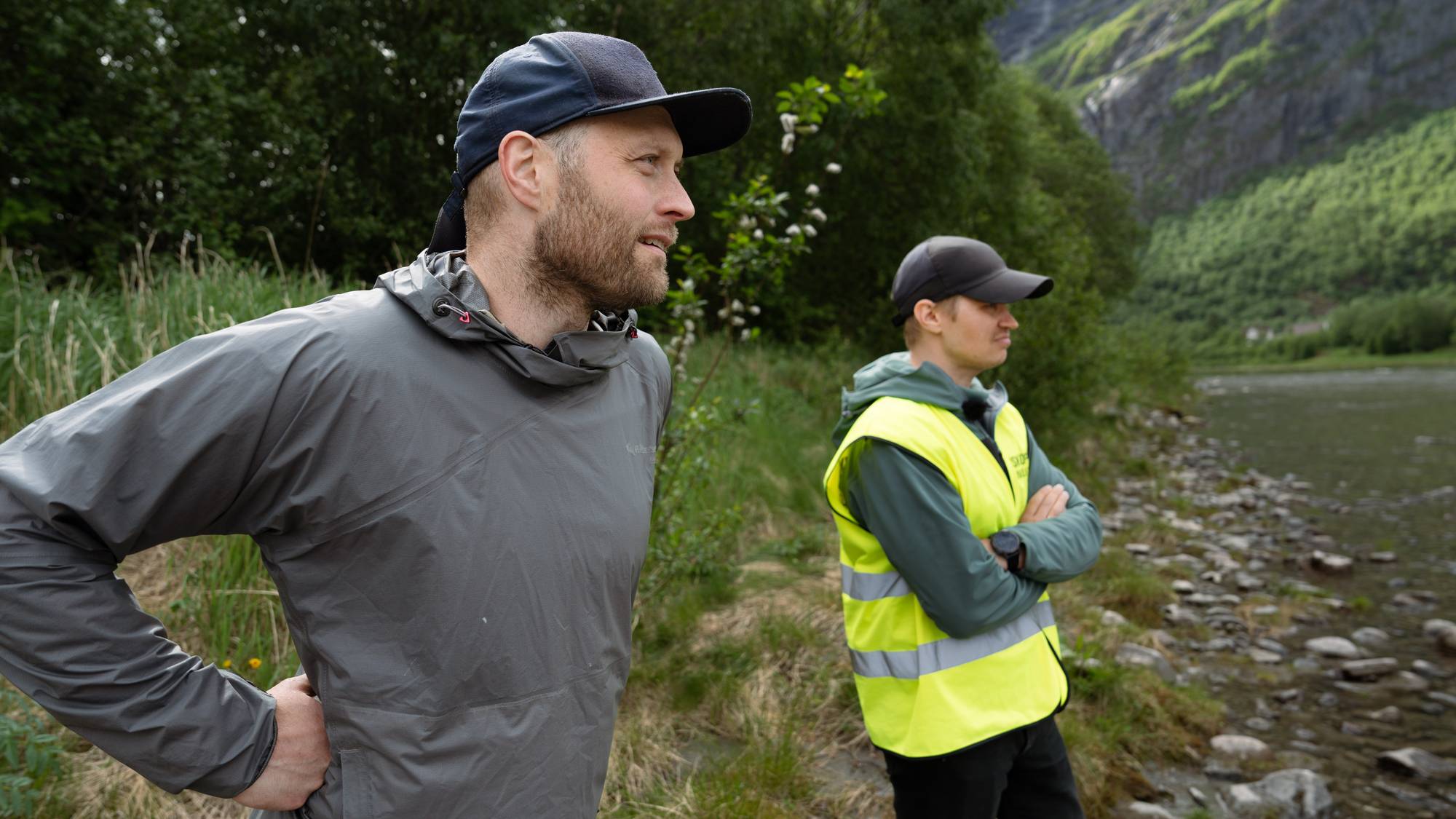 To menn står ved en elv i et fjellandskap. Den ene mannen, foran i bildet, har på seg en grå jakke og keps. Mannen bak har på seg en gul refleksvest og keps. (Bildebeskrivelsen er laget av en KI-tjeneste)