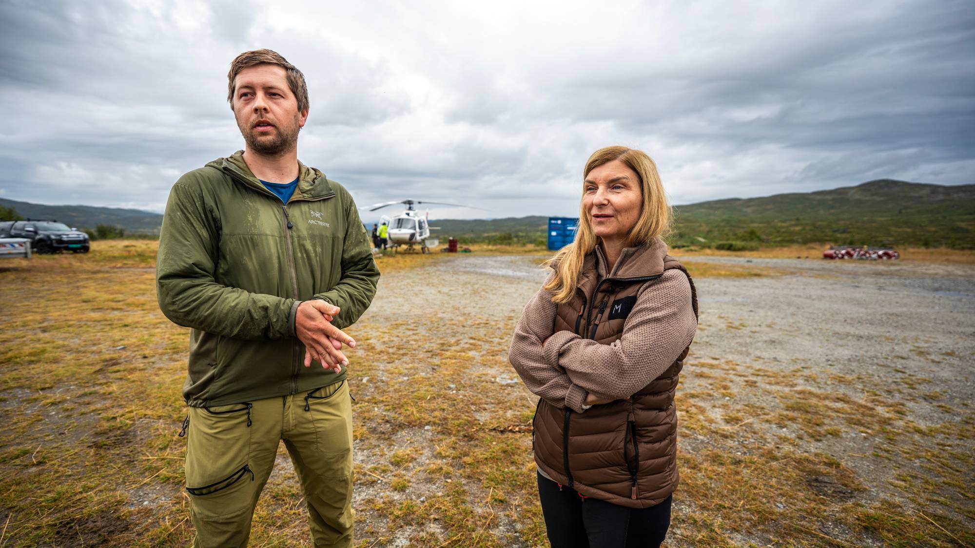 En mann og en kvinne står utendørs i et åpent landskap med et helikopter i bakgrunnen. (Bildebeskrivelsen er laget av en KI-tjeneste)