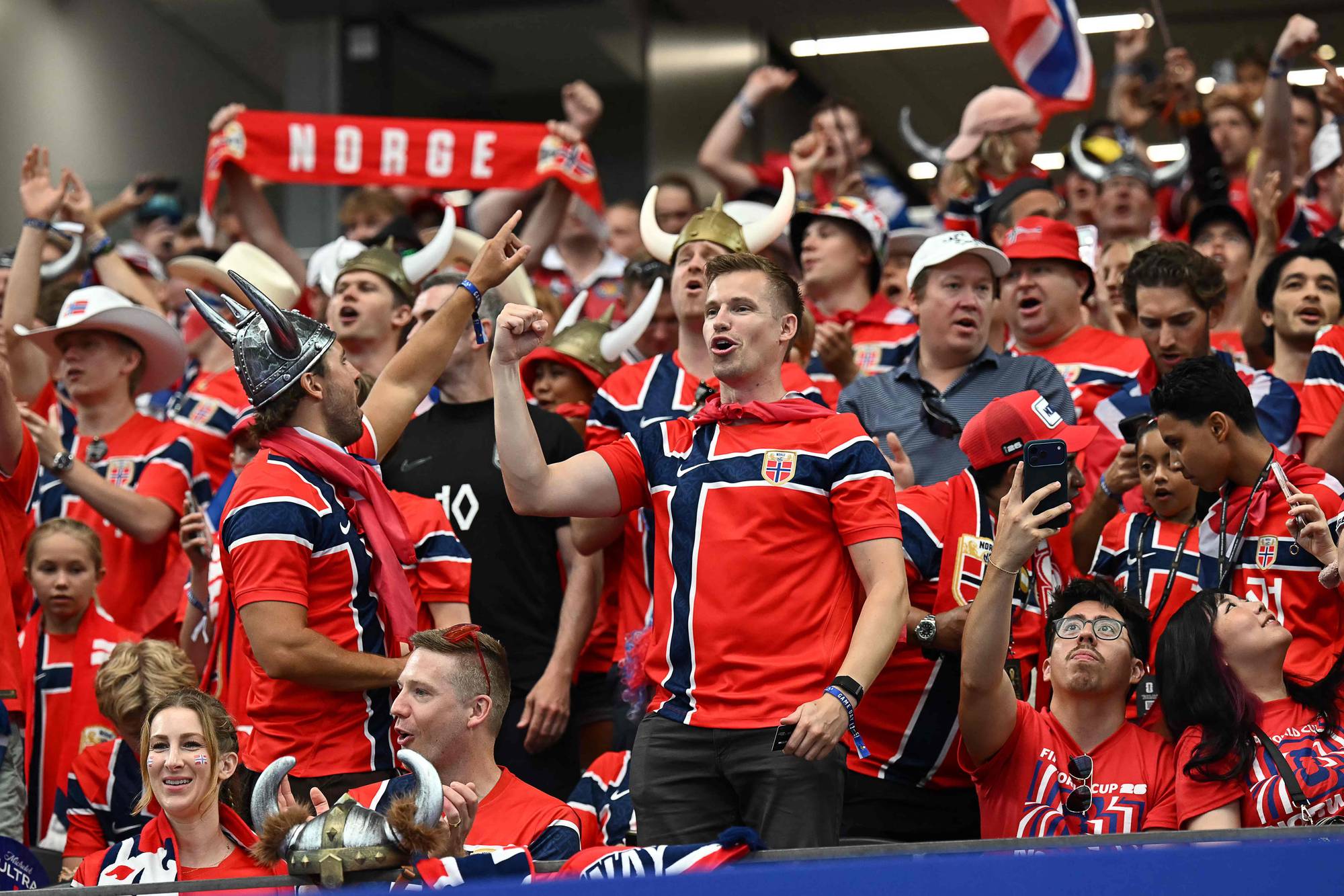 Norske supportere i landslagsdrakter heier på tribunen, mange med vikinghjelmer og flagg. (Bildebeskrivelsen er laget av en KI-tjeneste)