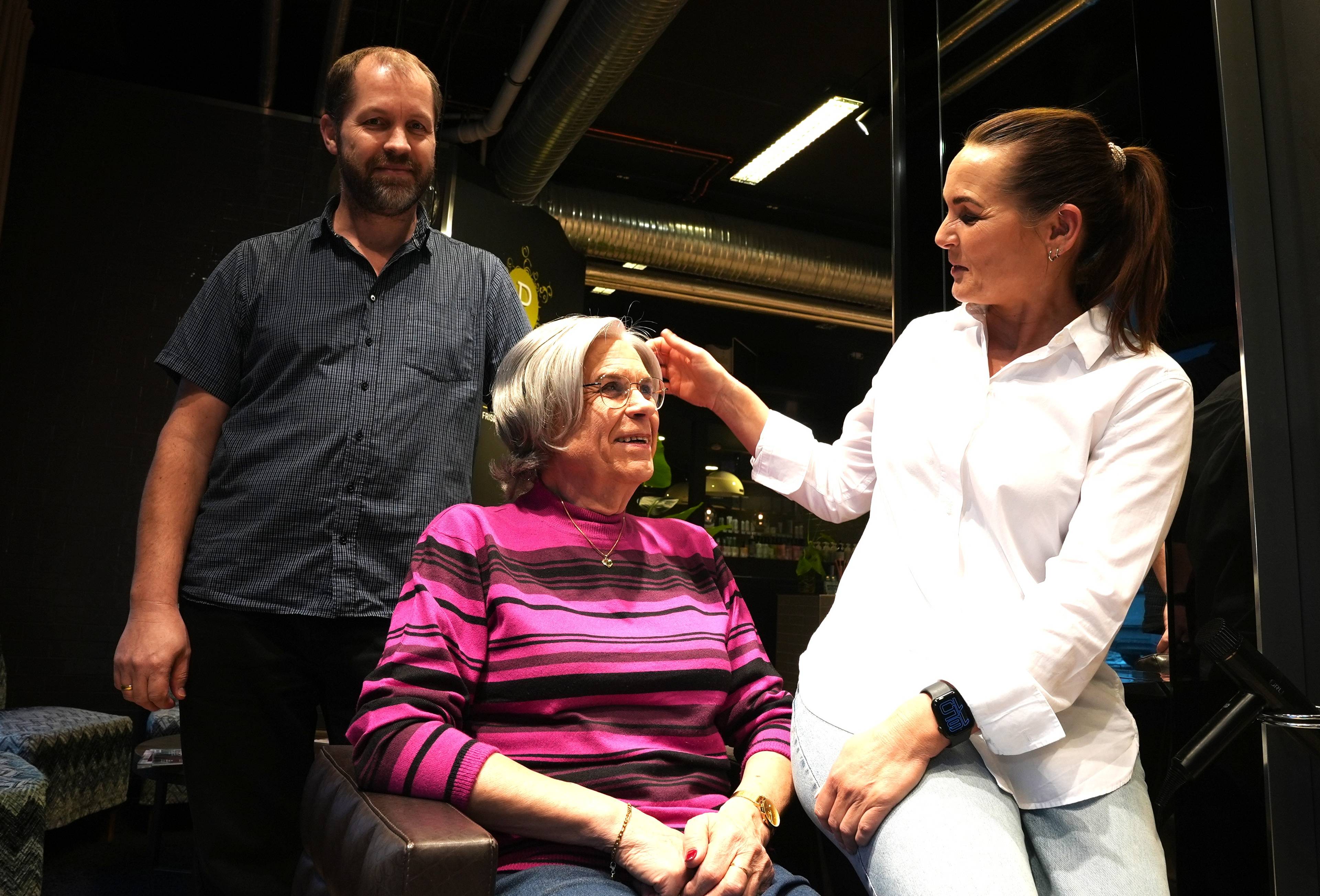 En mann står bak en eldre kvinne som sitter i en stol. En kvinne med hestehale justerer kvinnens hår. Kvinnen i stolen er kledd i en rosa og svart stripete genser. Mannen er kledd i en mørk skjorte. (Bildebeskrivelsen er laget av en KI-tjeneste)