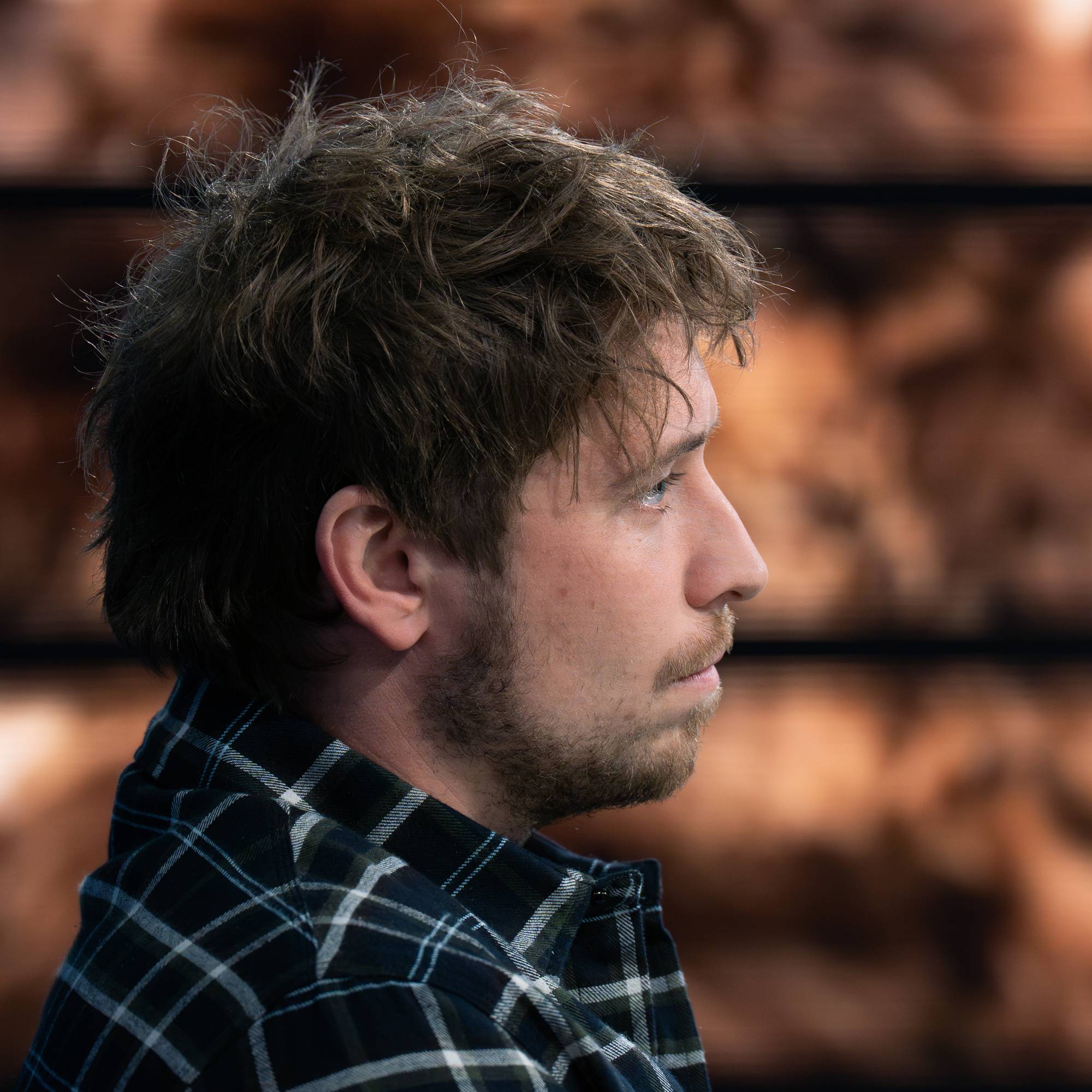 Profilbilde av en mann med brunt, rufsete hår. Han har på seg en rutete skjorte i mørke og lyse farger. Mannen ser til siden mens en bakgrunn med brede horisontale striper og uskarpe brune former er synlig. (Bildebeskrivelsen er laget av en KI-tjeneste)