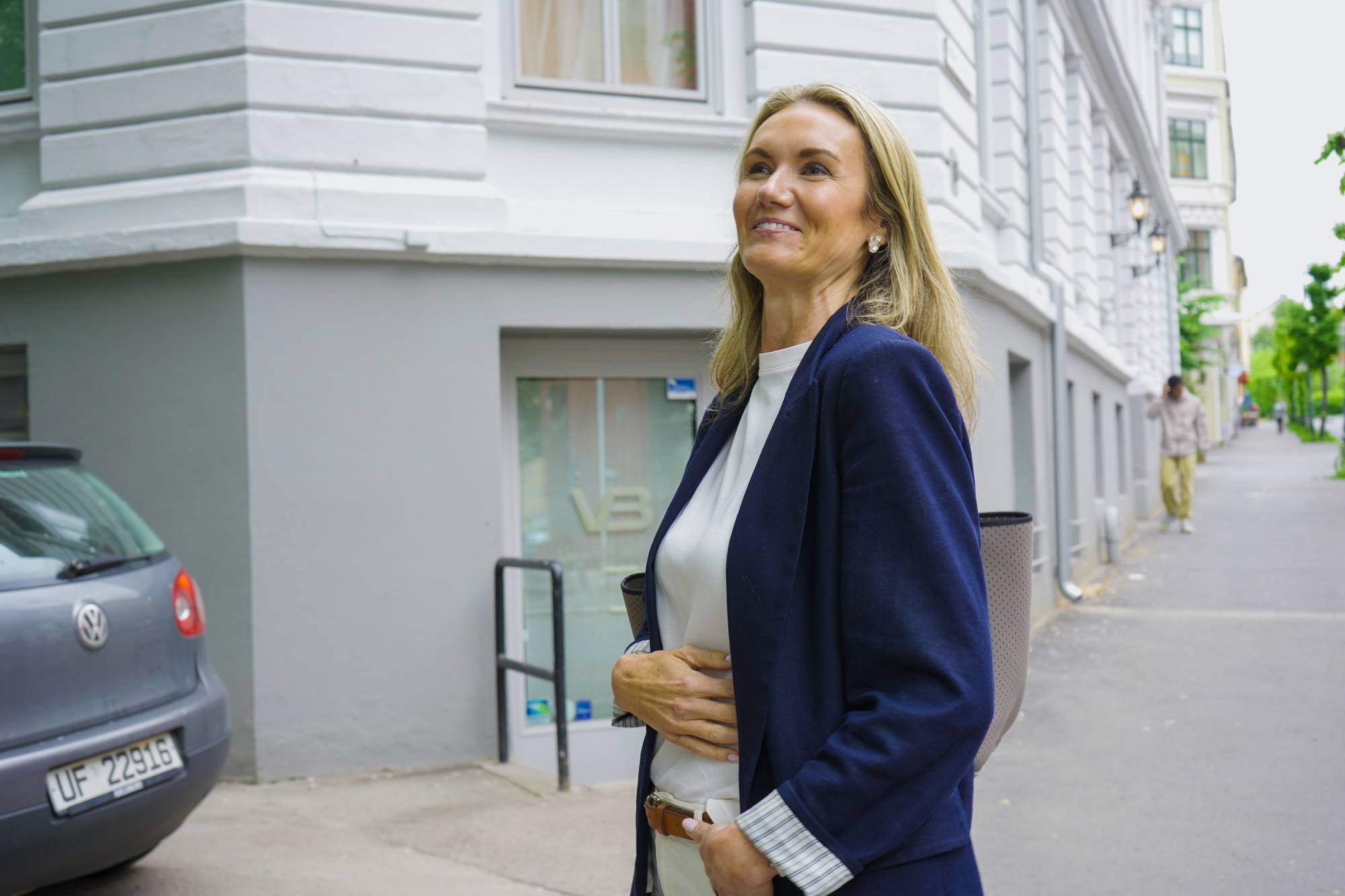 En kvinne i mørk blazer går ute. Bak henne står en bil parkert ved en bygning. En mann går på fortauet i bakgrunnen. (Bildebeskrivelsen er laget av en KI-tjeneste)