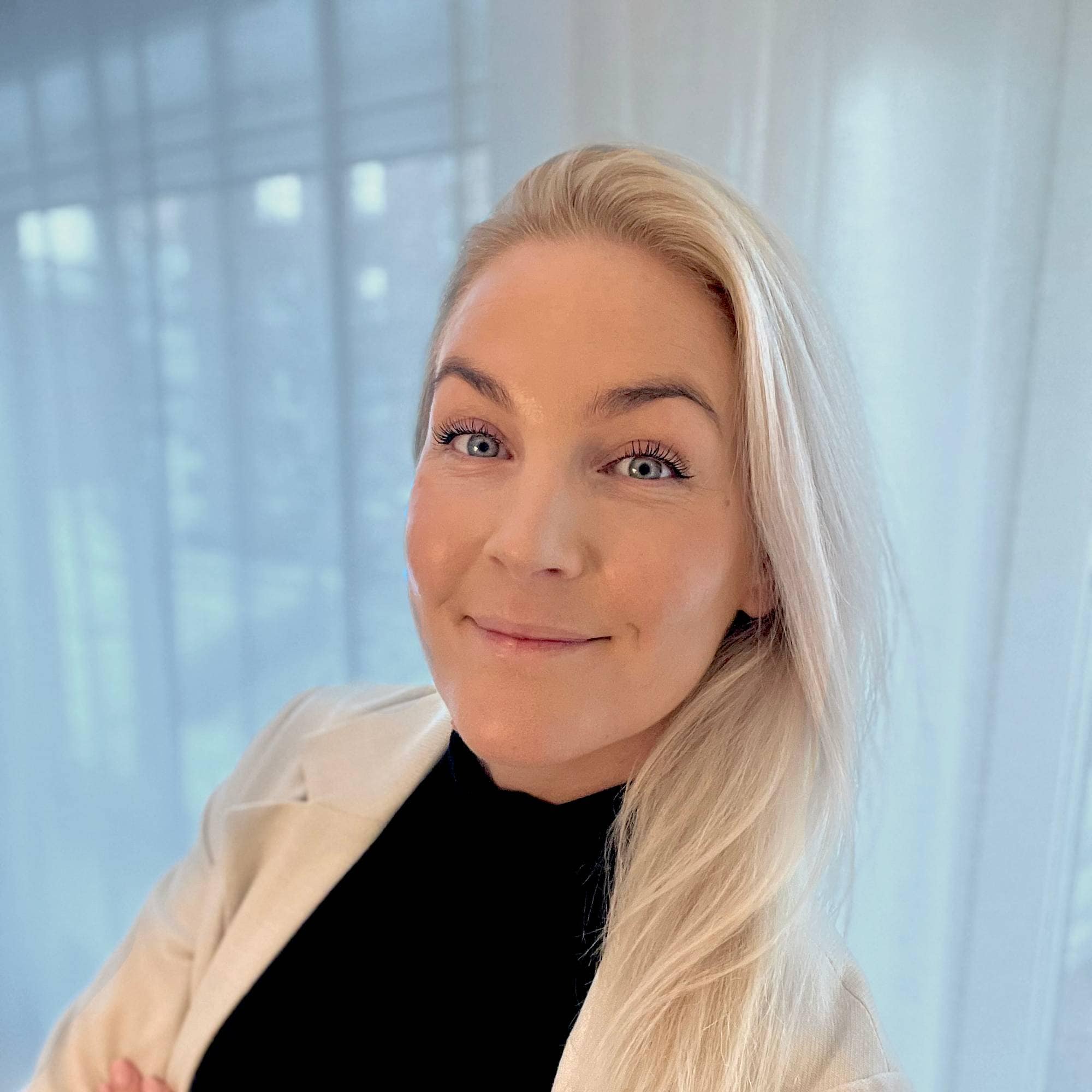 En kvinne med lyst, langt hår står foran en hvit gardin. Hun har på seg en svart t-skjorte og en lys jakke.