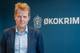 JON PETRUSSON / NRK En mann i mørk dress og lyseblå skjorte ser mot kamera. I bakgrunnen er et blått banner med et våpenskjold og teksten ØKOKRIM. (Bildebeskrivelsen er laget av en KI-tjeneste)