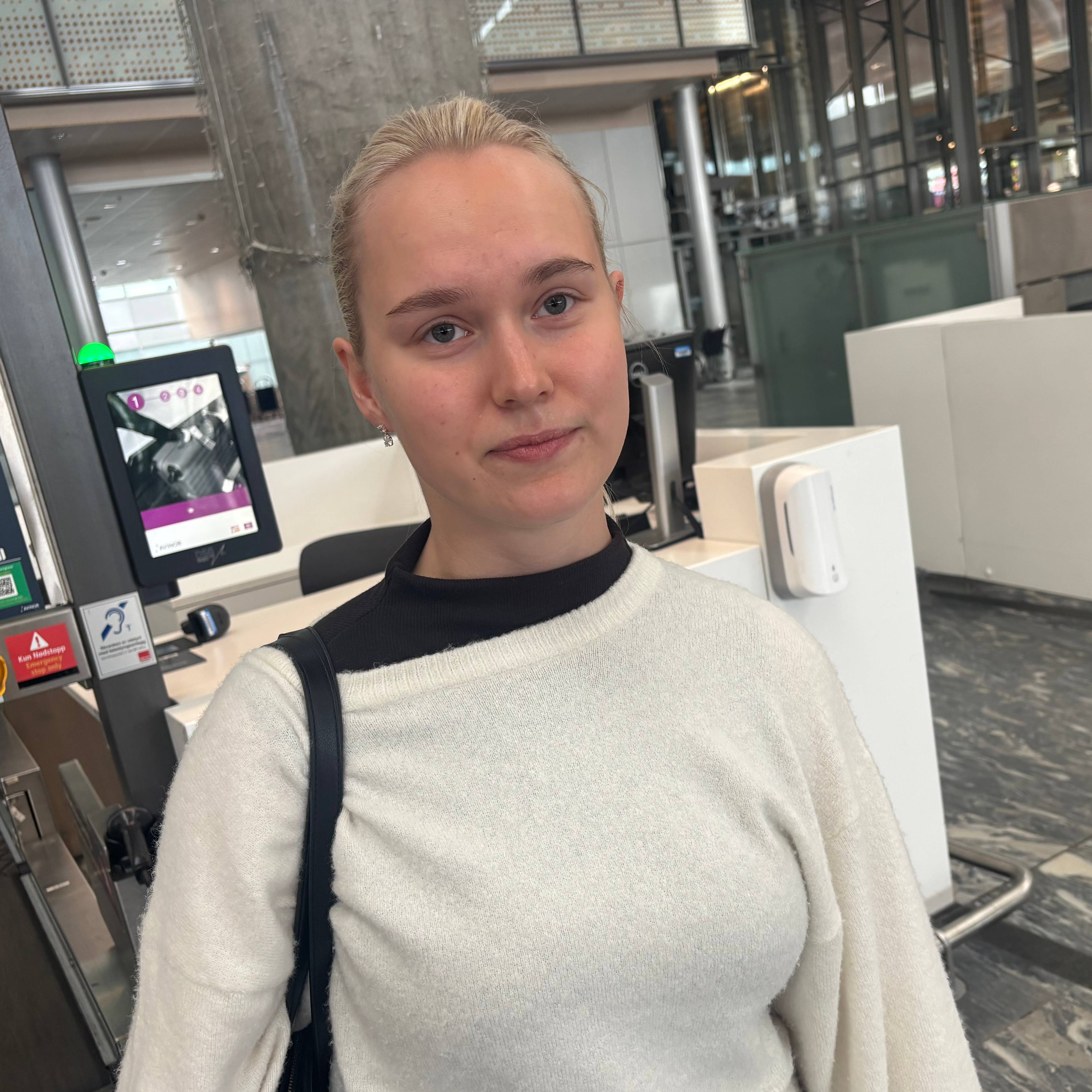 En kvinne står foran en skanner på en flyplass. Hun har på seg en hvit genser og har en veske over skulderen.