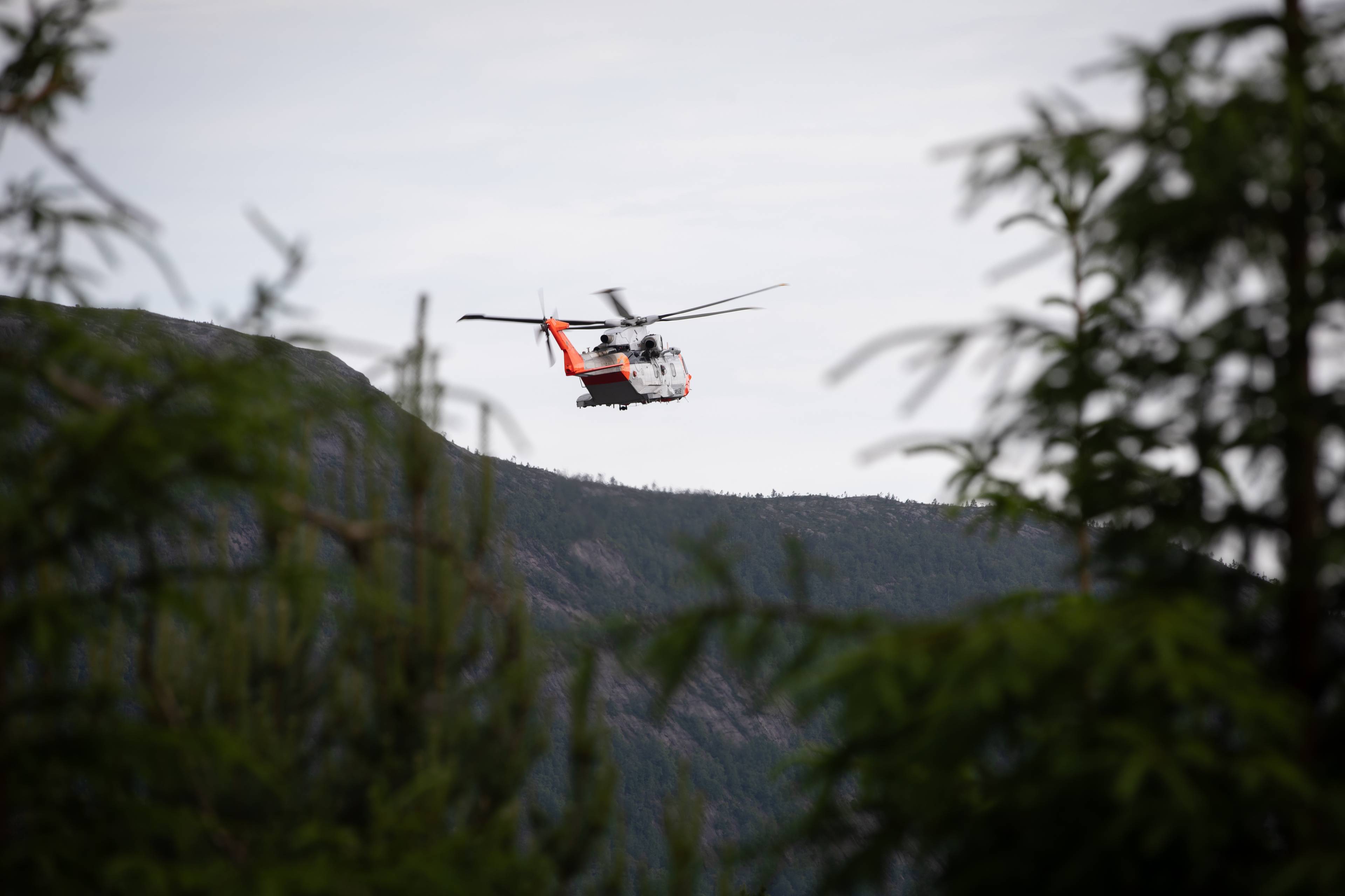 Et redningshelikopter flyr lavt over en åsside sett gjennom forgrunnen av trær. (Bildebeskrivelsen er laget av en KI-tjeneste)