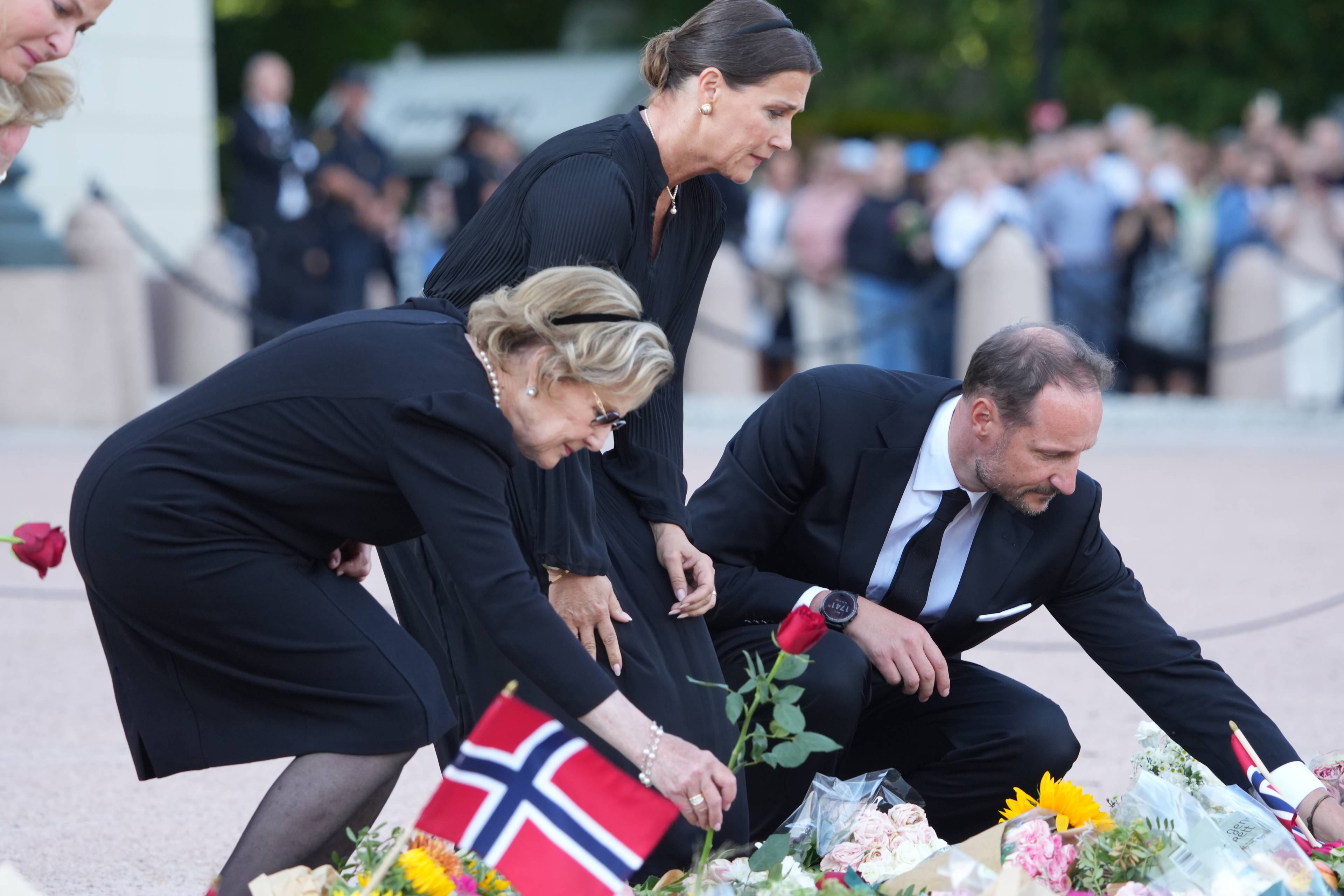 Dronning Sonja, prinsesse Mrtha Louise og kong Haakon legger ned roser foran Slottet. (Bildebeskrivelsen er laget av en KI-tjeneste)