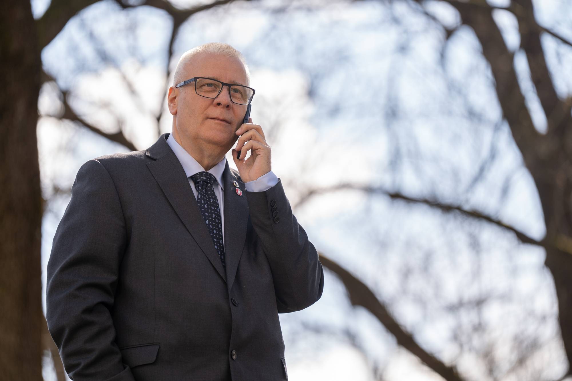 En mann i dress snakker i mobiltelefon. Han har briller og slips. Bakgrunnen er uskarpe trær og himmel. (Bildebeskrivelsen er laget av en KI-tjeneste)