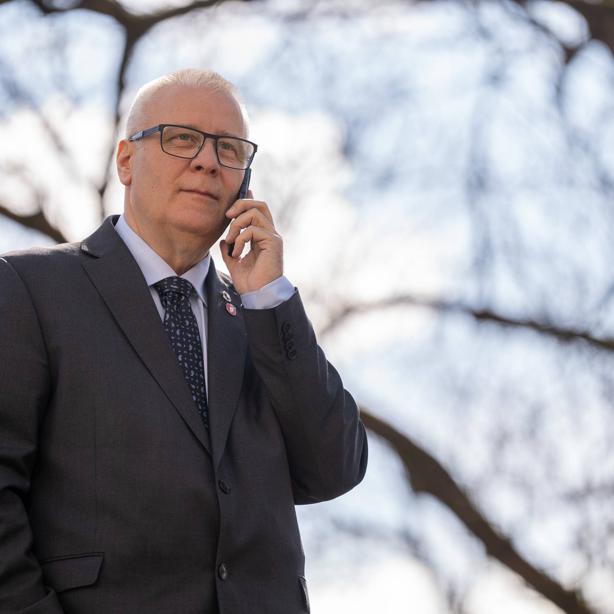 En mann i dress snakker i mobiltelefon. Han har briller og slips. Bakgrunnen er uskarpe trær og himmel. (Bildebeskrivelsen er laget av en KI-tjeneste)