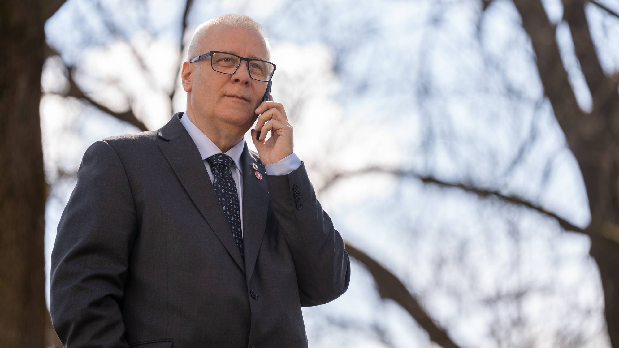 En mann i dress snakker i mobiltelefon. Han har briller og slips. Bakgrunnen er uskarpe trær og himmel. (Bildebeskrivelsen er laget av en KI-tjeneste)