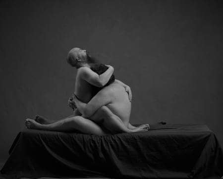 To nakne menn holder rundt hverandre på en seng med mørkt laken. Mannen uten hår, men med skjegg sitter på fanget til den andre, og holder den ene hånda rundt hodet hans. Den andre mannen med mørkt hår holder armene rundt han andre.