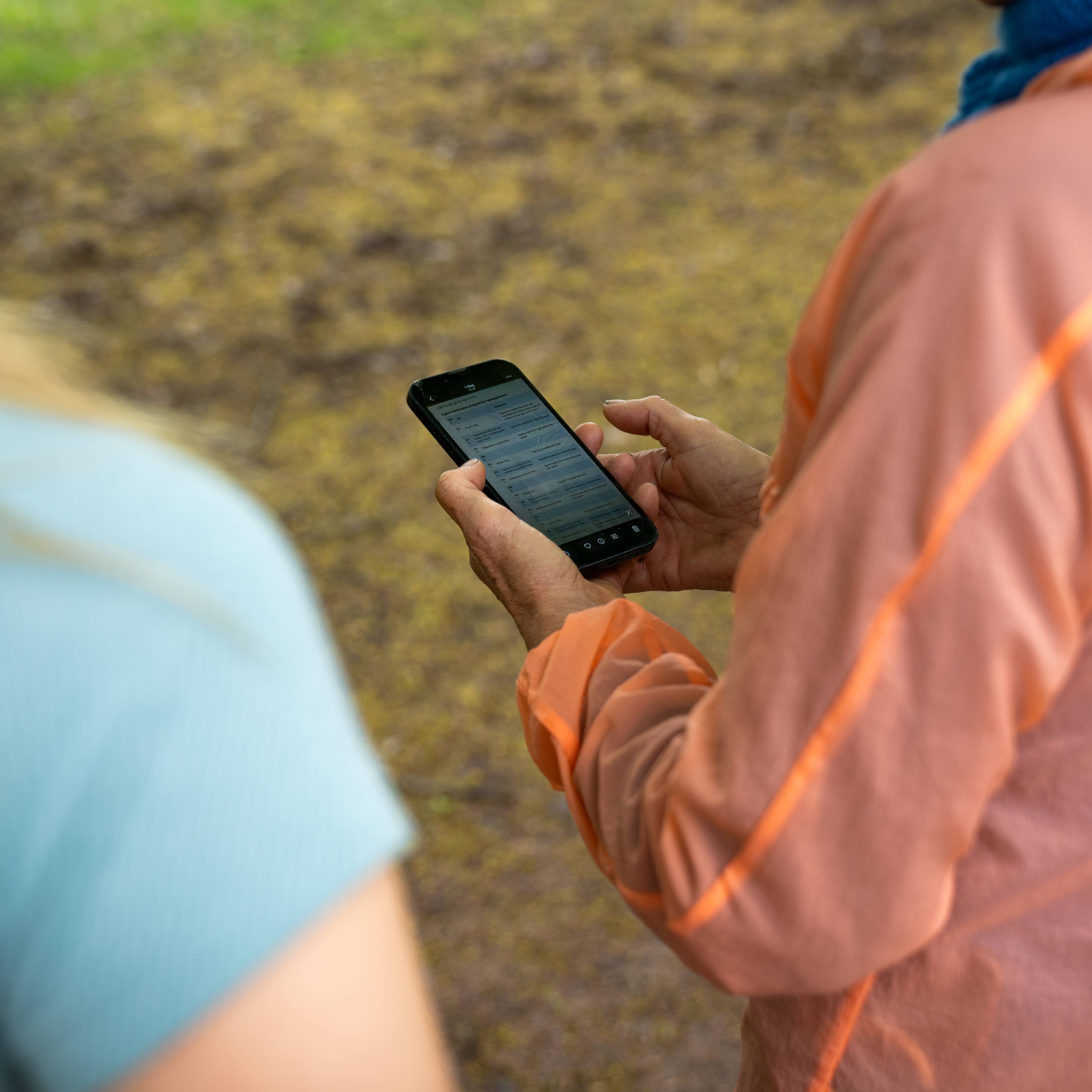 En kvinne holder en telefon. To personer ser på telefonen. Bildet er tatt utendørs i en park.