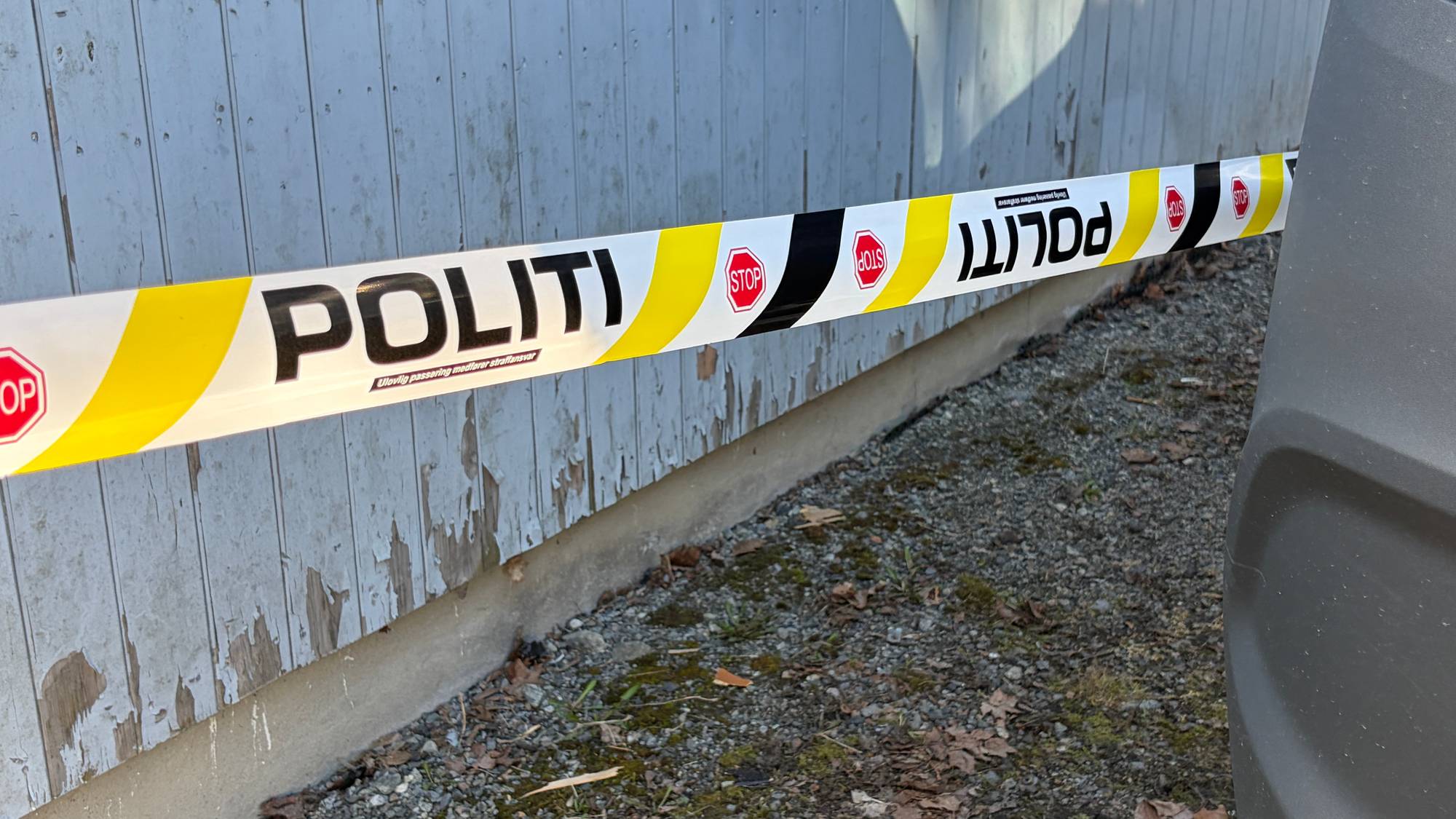 Sperrebånd med teksten POLITI er festet foran en falmet trevegg. Båndet har gule og svarte striper. En del av en bil er synlig til høyre. Rundt en søppelkasse er sperrebåndet festet, som er synlig lengre bak i bildet. (Bildebeskrivelsen er laget av en KI-tjeneste)