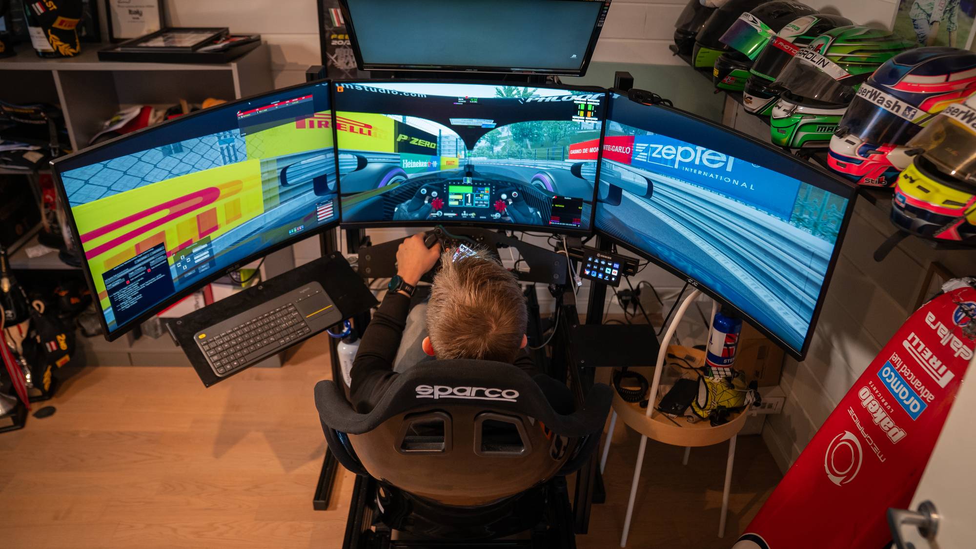 En person sitter i en racingstol foran et trippel-skjermoppsett og spiller et racingspill. Flere hjelmer er stablet på en hylle til høyre. Det er et lite skrivebord med tastatur foran skjermene. En stor rød gjenstand med sponsornavn er lent mot veggen. (Bildebeskrivelsen er laget av en KI-tjeneste)