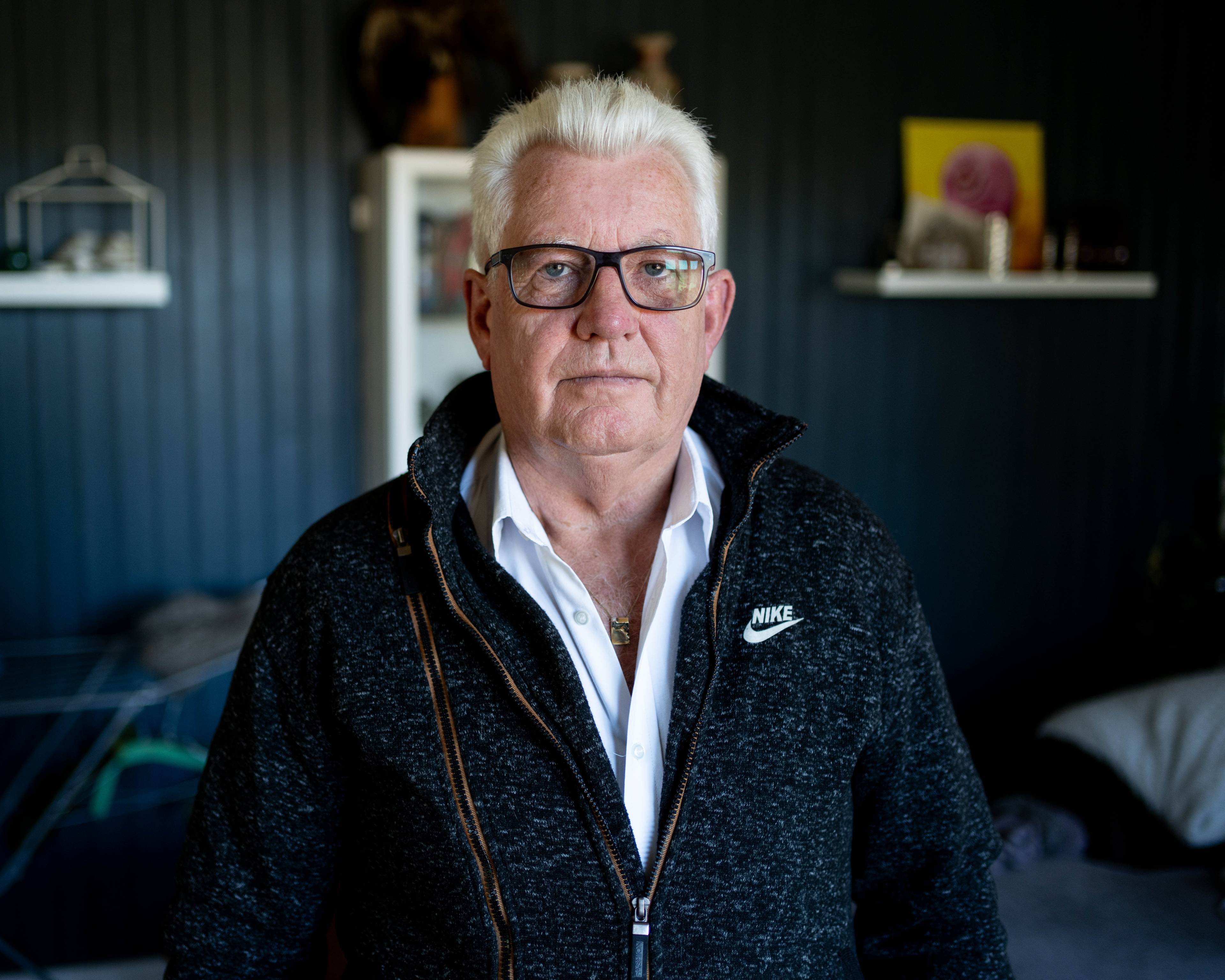 Erik Arntzen  ser rett i kamera. han har på seg flis og hvit skjorte, han har briller og hvitt hår. Han står i en stue.