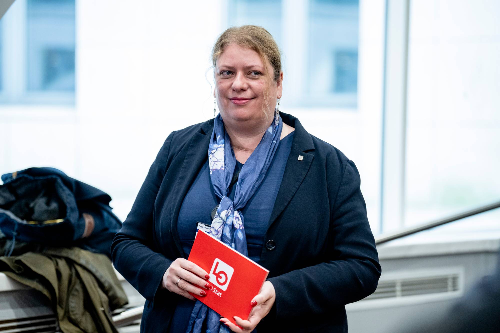 En kvinne med blondt hår, iført mørk dress og blått skjerf, holder en rød brosjyre. Kvinnen har et smil om munnen. Bak henne er det stablet klær. Bildet er tatt innendørs. (Bildebeskrivelsen er laget av en KI-tjeneste)