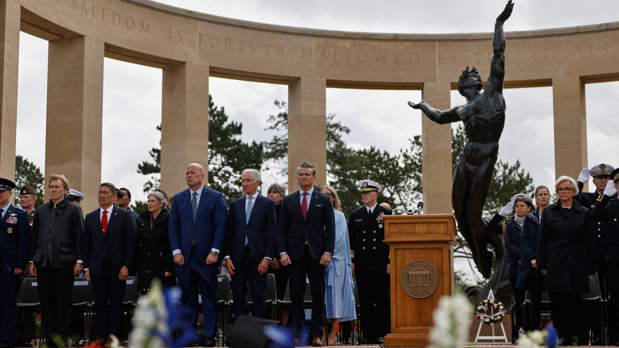 En gruppe personer, inkludert militært personell og sivile, står samlet foran et monument og en statue. De fleste er kledd i formelle antrekk som dresser og uniformer. Bak dem er en stor, buet struktur med inskripsjon. Det er blomsterdekorasjoner foran scenen. (Bildebeskrivelsen er laget av en KI-tjeneste)