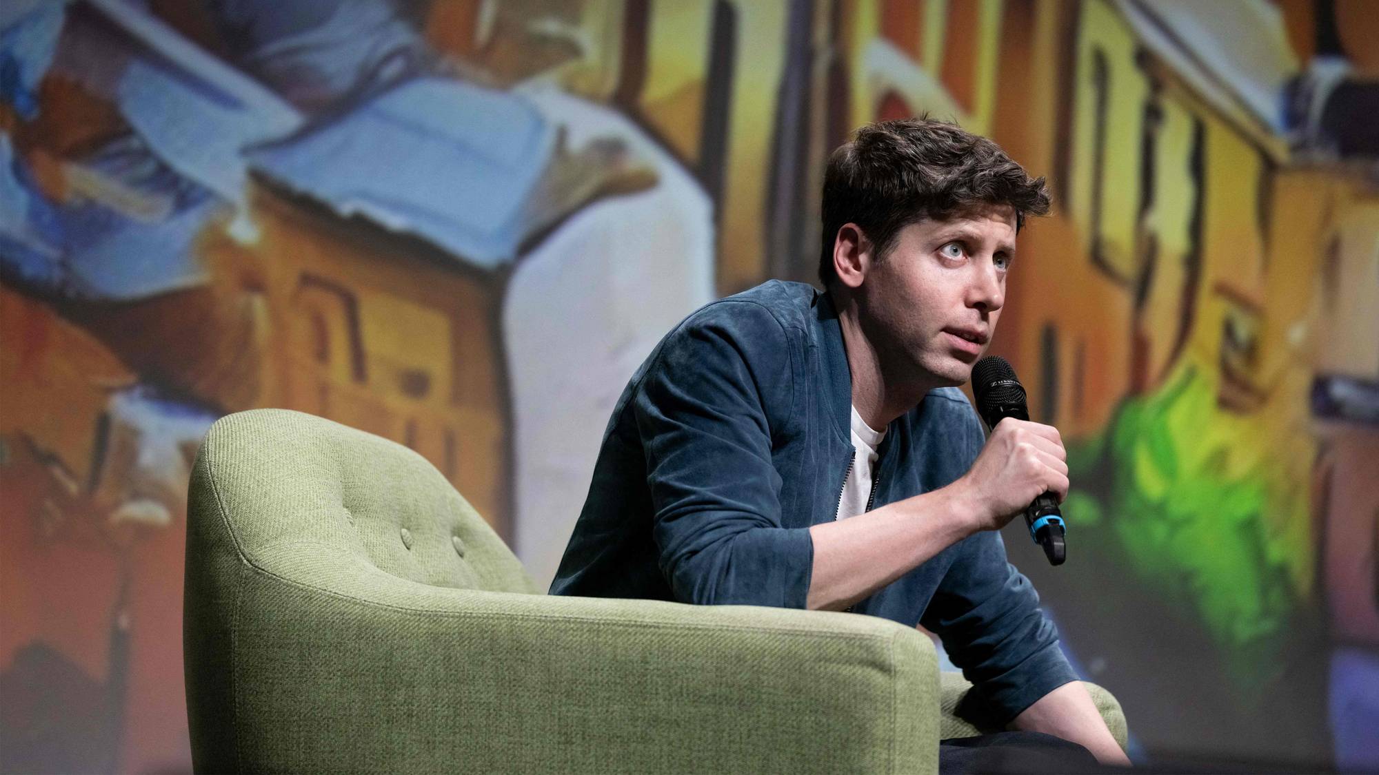 Bilde av Sam Altman sjefen i OpenAI. Han sitter på en scene med en mikrofon i hånden. 