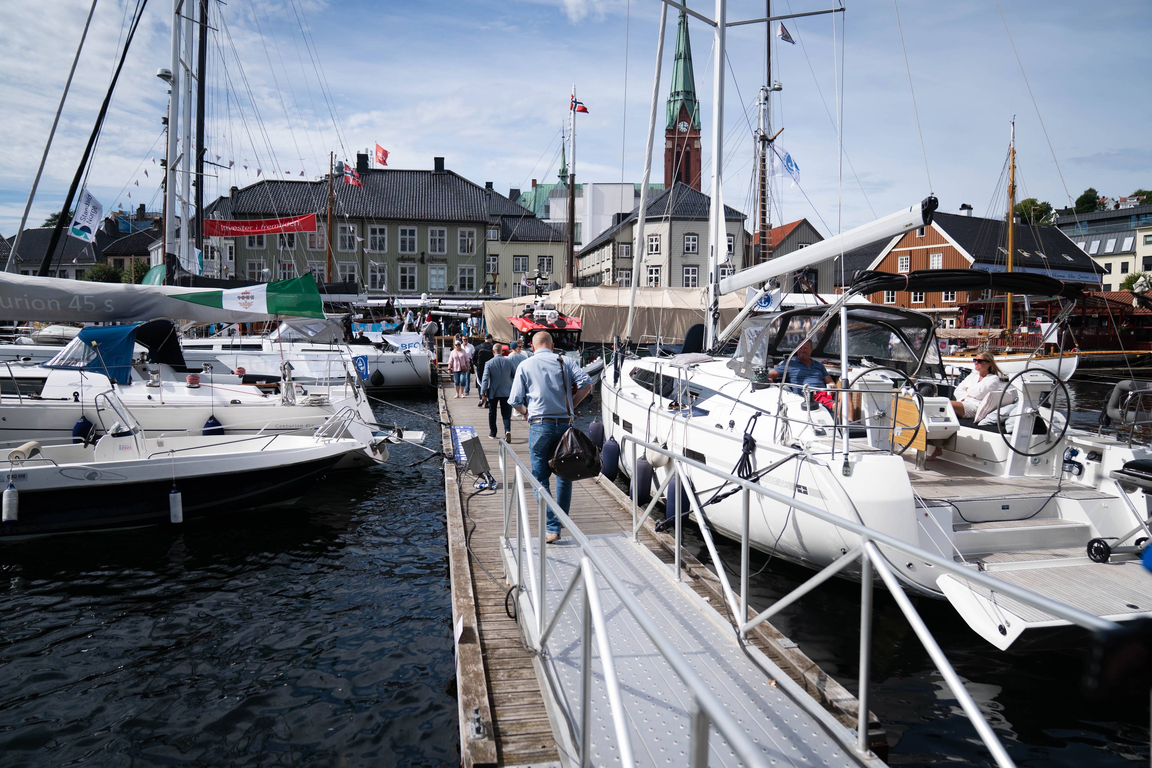 Personer går på en brygge omgitt av seilbåter i en havn. I bakgrunnen ses eldre trehus og en kirke. (Bildebeskrivelsen er laget av en KI-tjeneste)