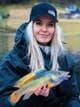 Tale Hauso En kvinne med blondt hår og en svart caps holder en fargerik fisk. Hun er kledd i en mørk jakke, og har på seg en vest over jakken. Bak henne sees et vann og bebyggelse i en fjellside. Et fiskestang er synlig i forgrunnen. (Bildebeskrivelsen er laget av en KI-tjeneste)