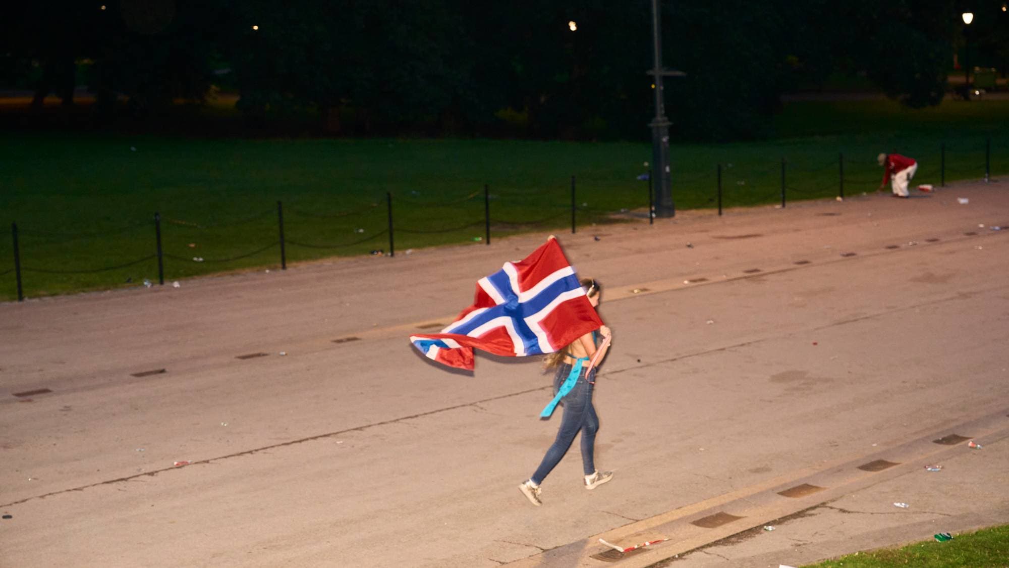 Person med norsk flagg går over en asfaltert flate ved en park om kvelden. 