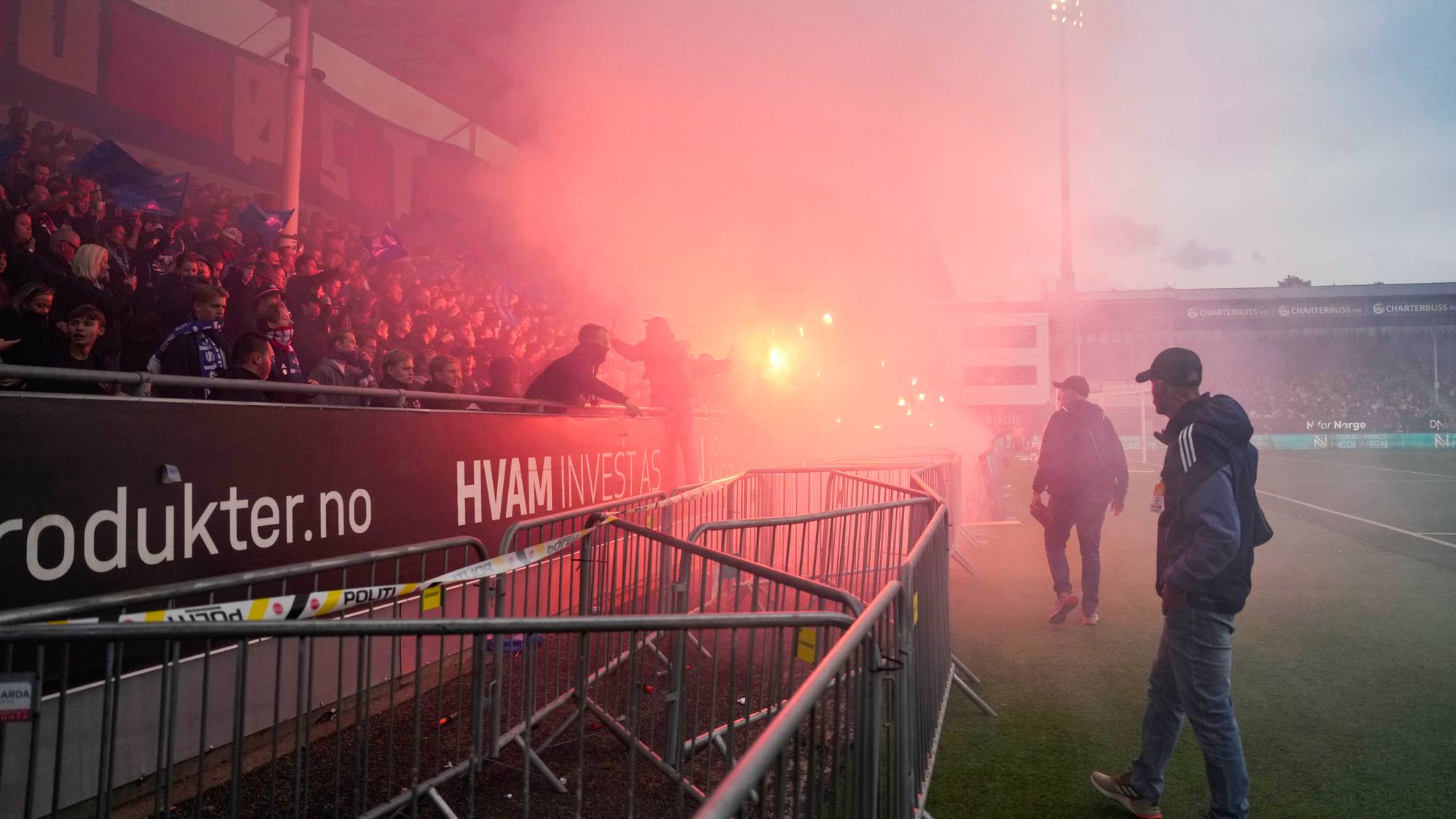 Supportere på en fotballtribune tenner røykbluss, som fyller deler av stadion med rød røyk. (Bildebeskrivelsen er laget av en KI-tjeneste)