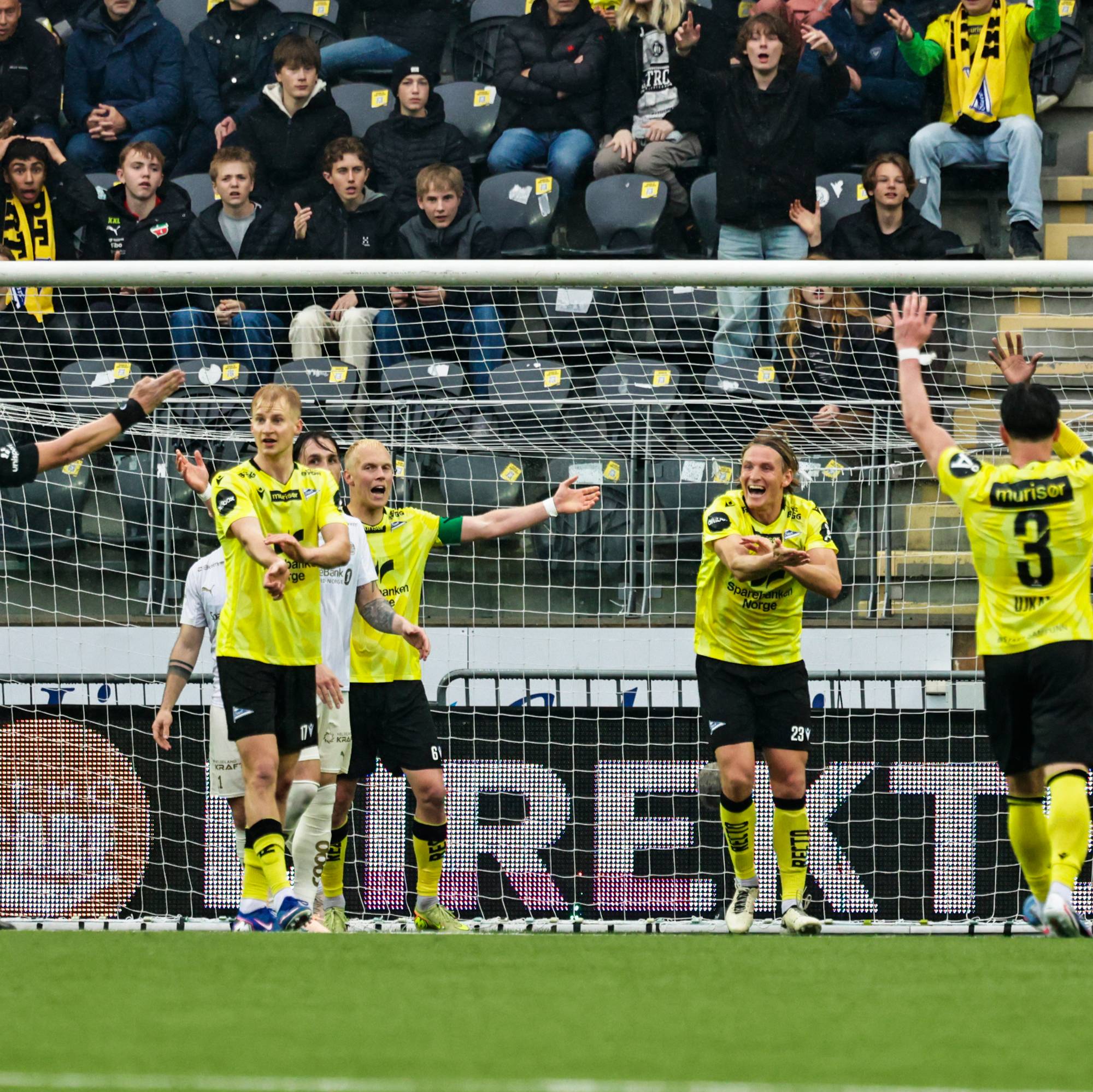 Fotballspillere i gule drakter feirer ved målet. En dommer i svart er til stede. Tilskuere sitter på tribunen bak dem. Spillerne ser ut til å ha scoret. (Bildebeskrivelsen er laget av en KI-tjeneste)