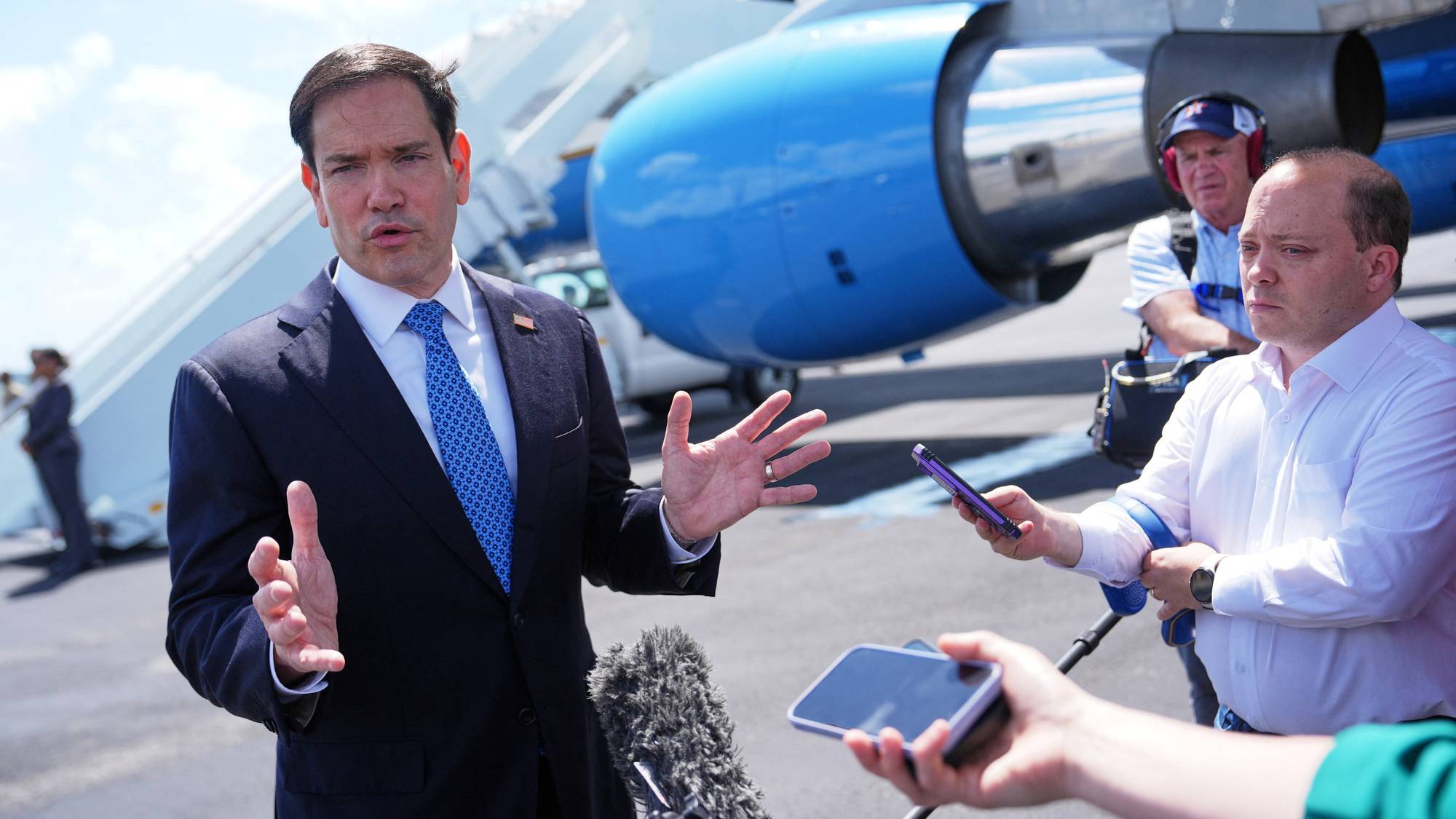 Politiker Marco Rubio snakker til media. En person holder ut en mobiltelefon for å ta opp uttalelser. I bakgrunnen er det et fly og en flytrapp. (Bildebeskrivelsen er laget av en KI-tjeneste)