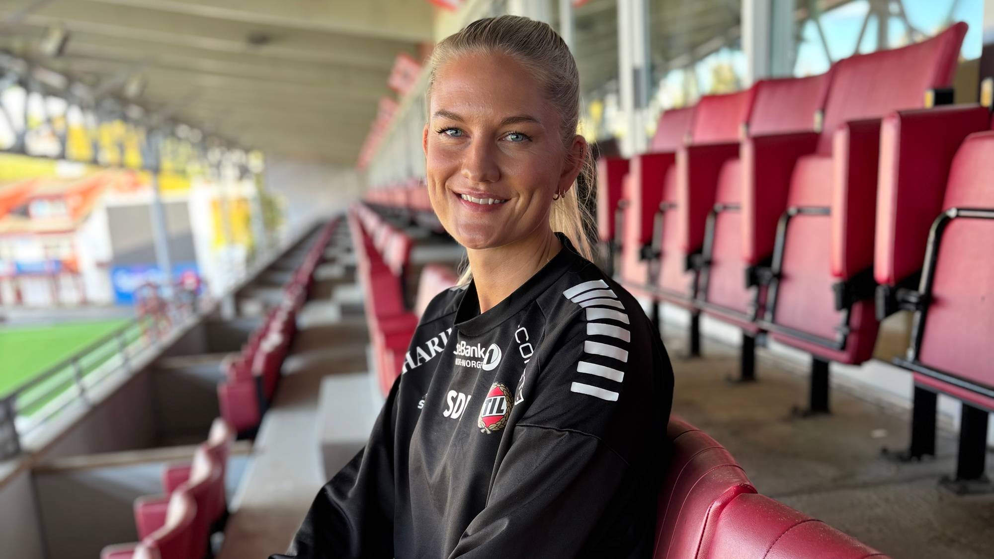 En kvinne i sportstøy ser i kamera og smiler. Sitter på en sete på en tribune.
