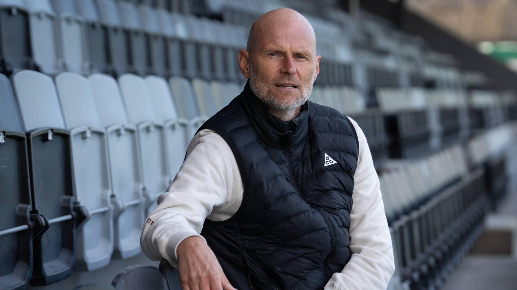 En mann sitter på en tribun på en stadion. Han har på seg en svart vest over en lys genser. Mannen ser inn i kameraet. Bak ham er det tomme seterader. (Bildebeskrivelsen er laget av en KI-tjeneste)