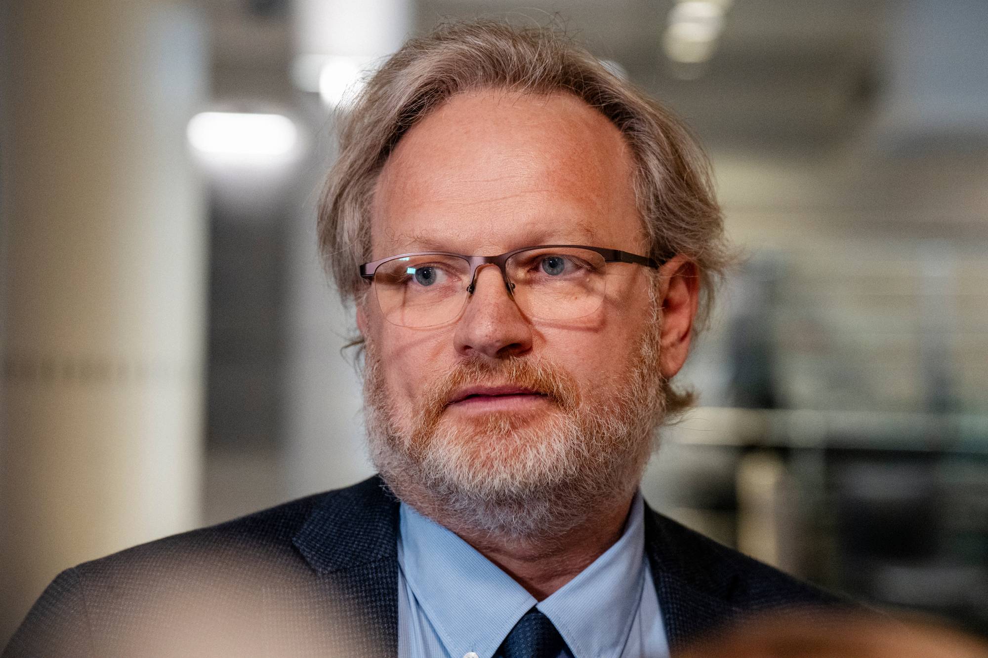 Et nærbilde av en mann med briller og skjegg. Mannen har grått hår og er iført dressjakke og skjorte. Bakgrunnen er uskarp og viser en innendørs setting med lys. (Bildebeskrivelsen er laget av en KI-tjeneste)