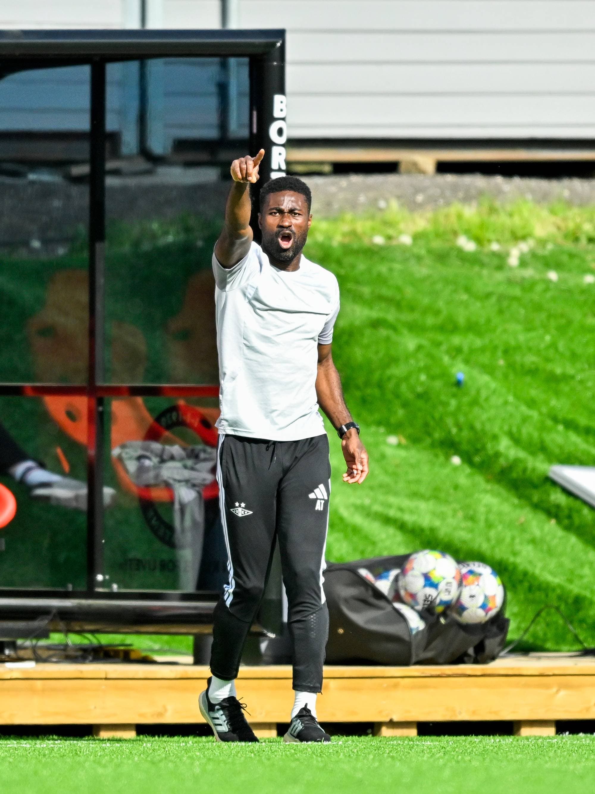 En fotballtrener peker og roper under en kamp på en gressbane. Ved siden av ham sitter en annen person foran en bærbar datamaskin. I bakgrunnen er en sportsbenk og baller i en bag. (Bildebeskrivelsen er laget av en KI-tjeneste)