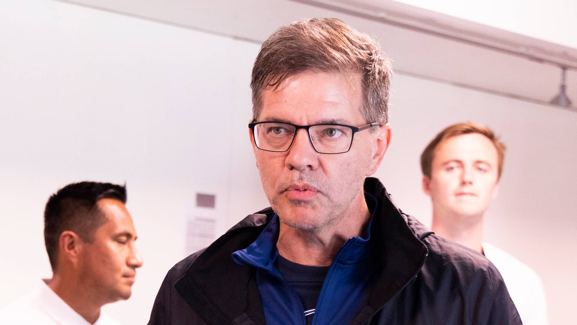Tre menn i sportsklær står innendørs, der en mann med briller snakker til de andre. (Bildebeskrivelsen er laget av en KI-tjeneste)