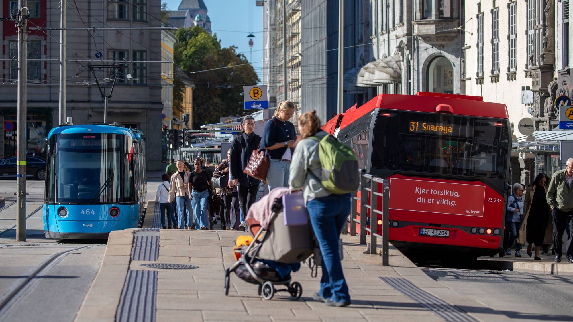Fotgjengere ved et busstopp i Oslo sentrum med en trikk og en buss i bakgrunnen. (Bildebeskrivelsen er laget av en KI-tjeneste)