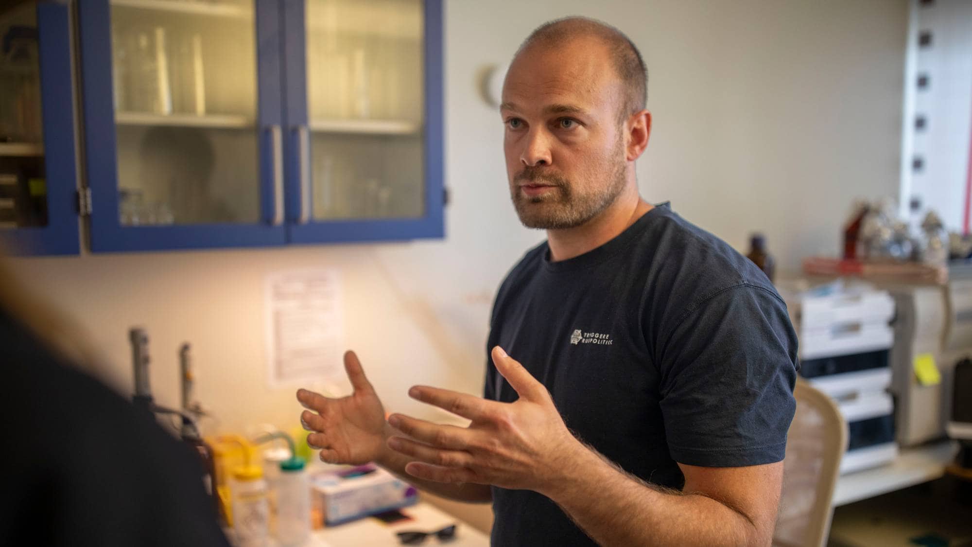 En mann i mørk t-skjorte snakker og gestikulerer med hendene. Bak ham er det et laboratorie eller et kontor med skap, hyller og utstyr. (Bildebeskrivelsen er laget av en KI-tjeneste)