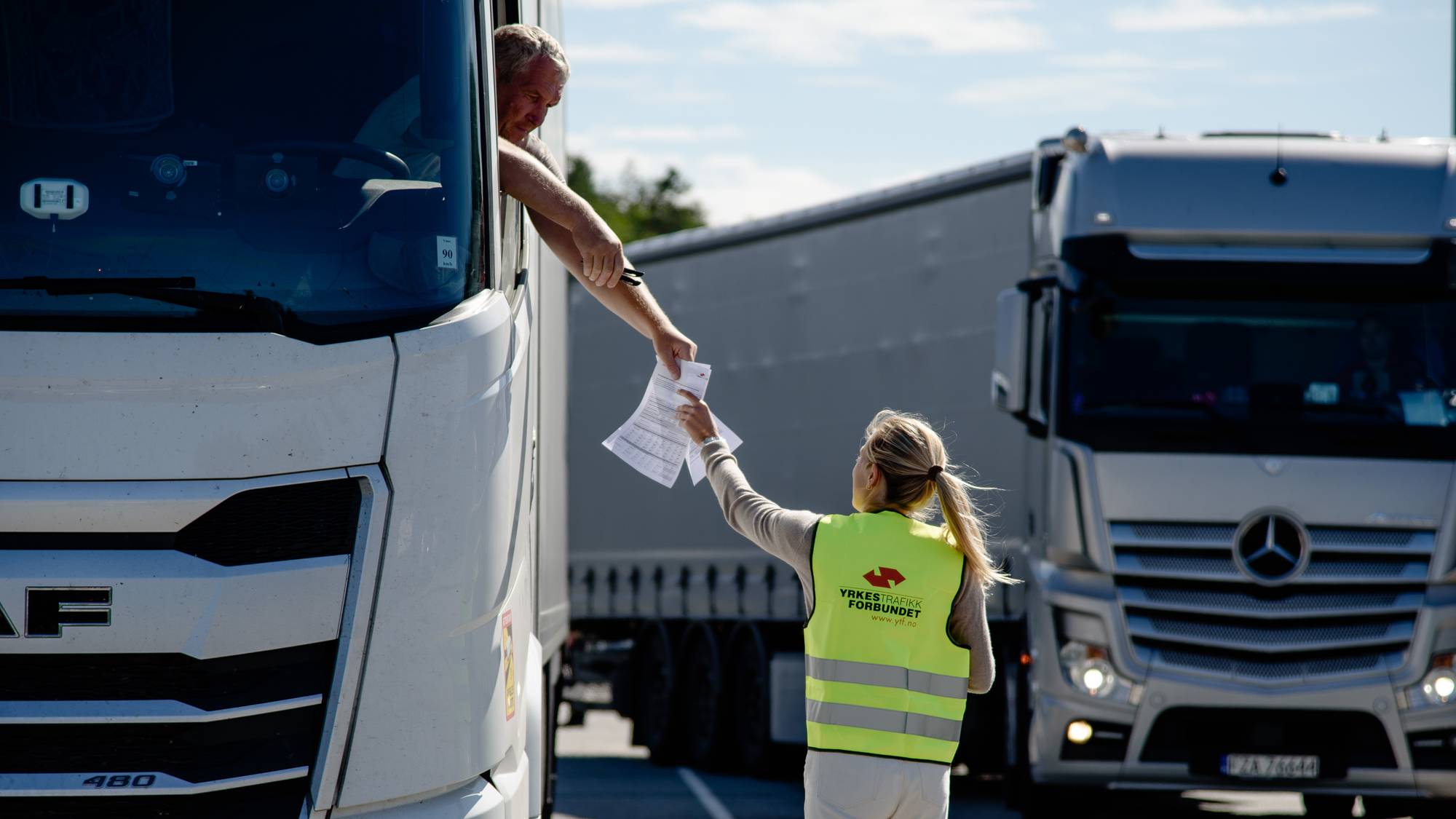 To hvite lastebiler står på en asfaltert plass. VEd den ene står en kvinen i gul vest med ryggen til, hun rekker et hvitt ark til en lastebilsjåfør som strekker hode og arm ut av vinduet. 