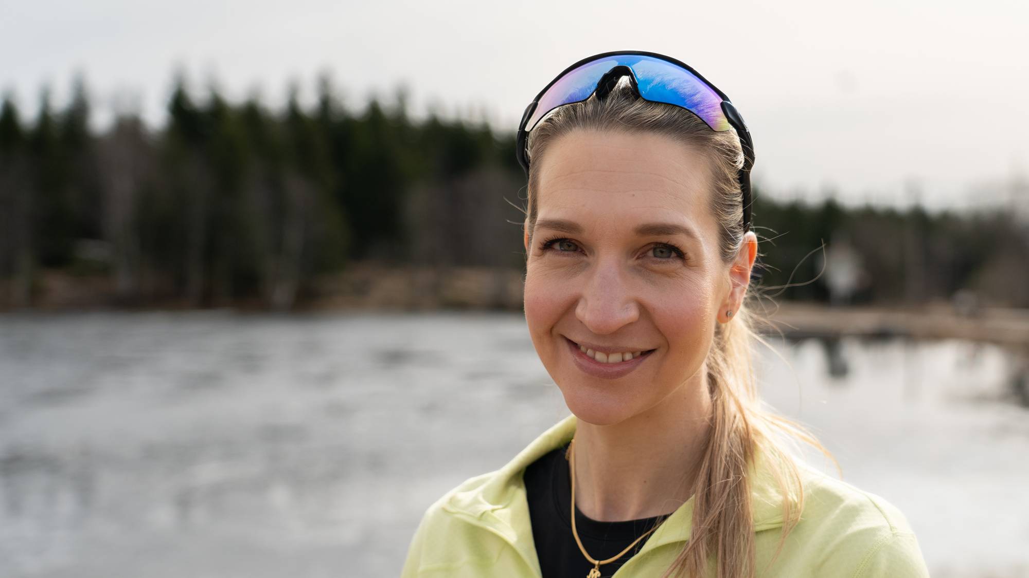 En kvinne med solbriller på hodet smiler mot kameraet. Hun er iført en lys grønn treningsoverdel. I bakgrunnen er det en innsjø og trær. (Bildebeskrivelsen er laget av en KI-tjeneste)
