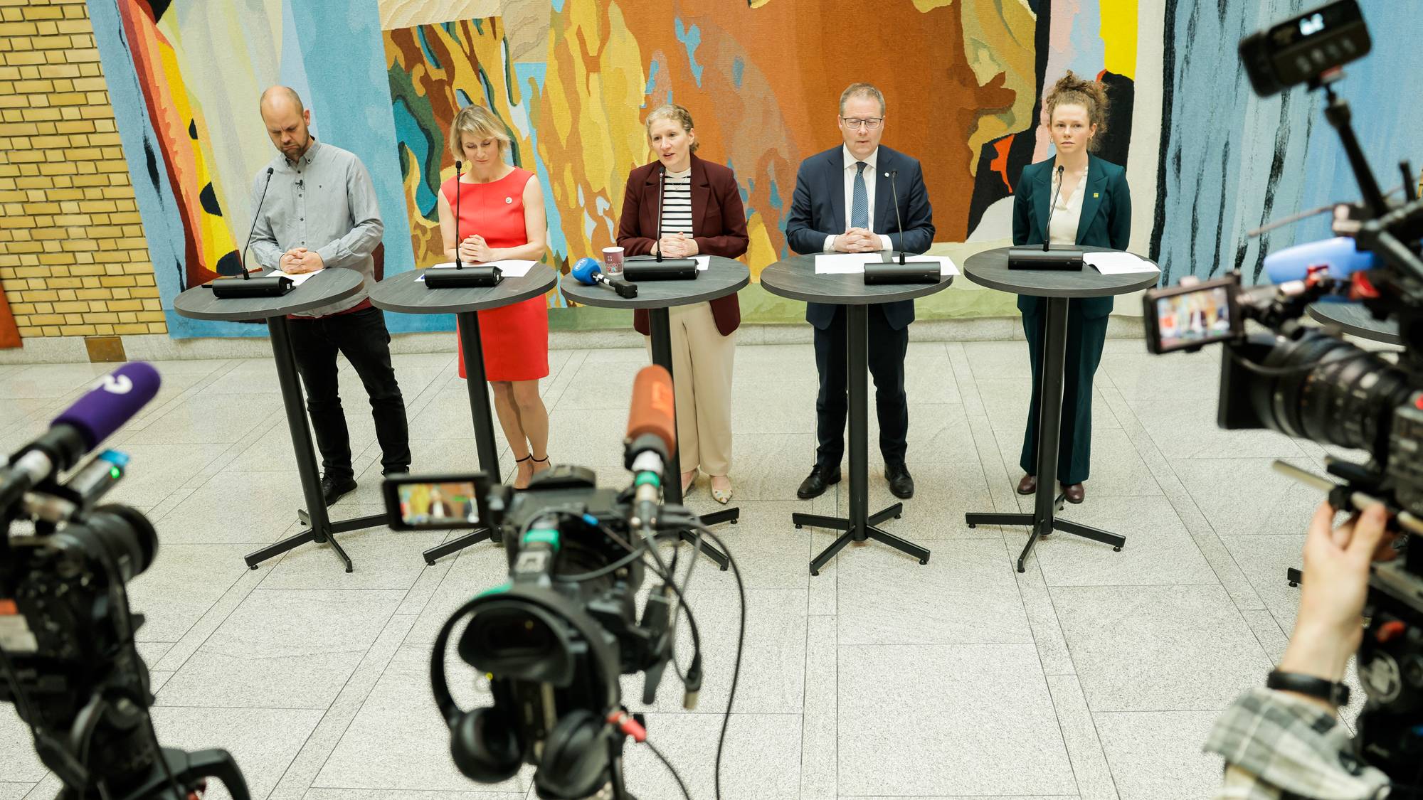 Fem personer står bak podier foran en stor billedvev. Det er flere kameraer og mikrofoner rettet mot personene. En person er i ferd med å justere et kamera. (Bildebeskrivelsen er laget av en KI-tjeneste)