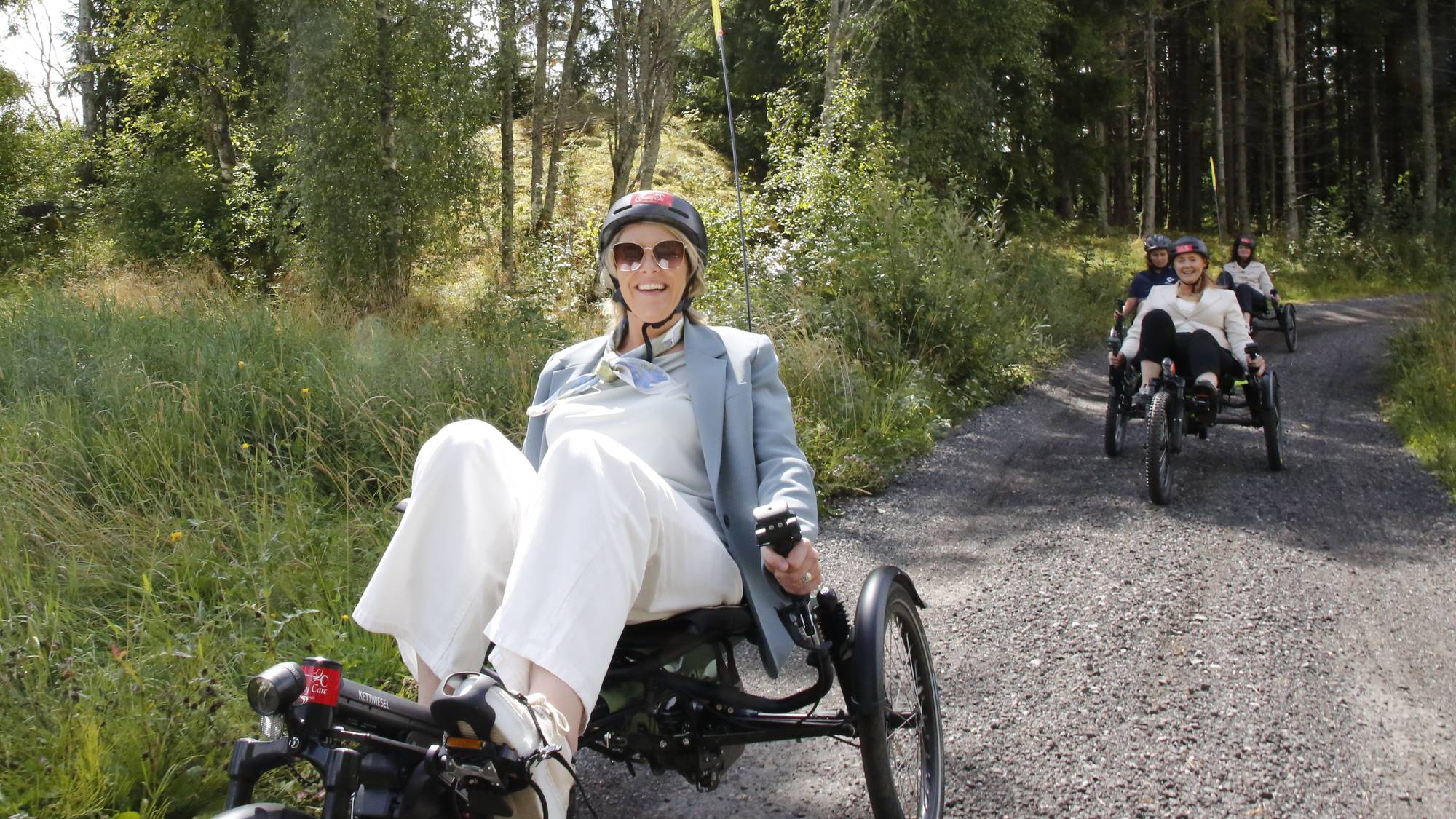 Sylvi Listhaug sykler på en liggesykkel på en grusvei i et skogsområde sammen med andre. 