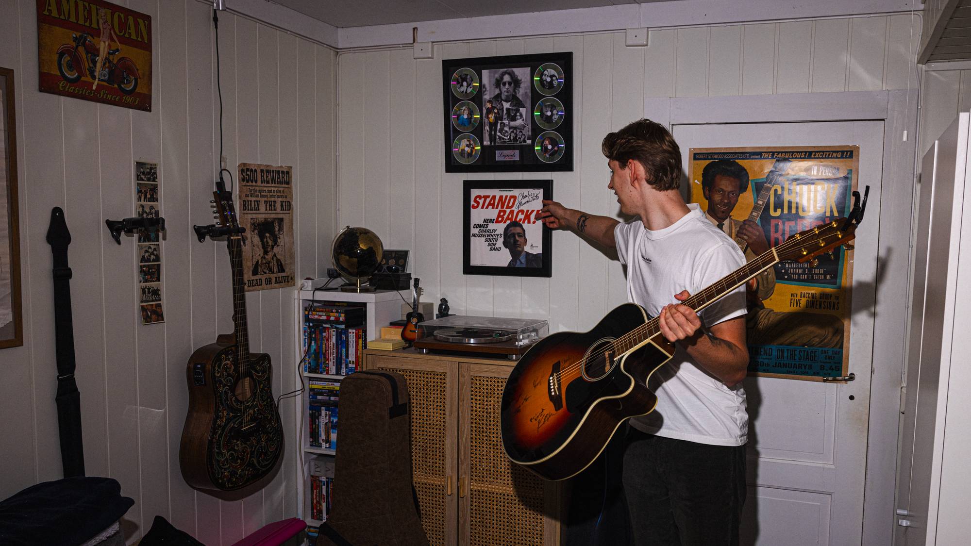 Foto av Trym inne på soverommet sitt.  Han holder en gitar i hånden. I bakgrunnen ser du en platespiller og flere plakater på veggene. 