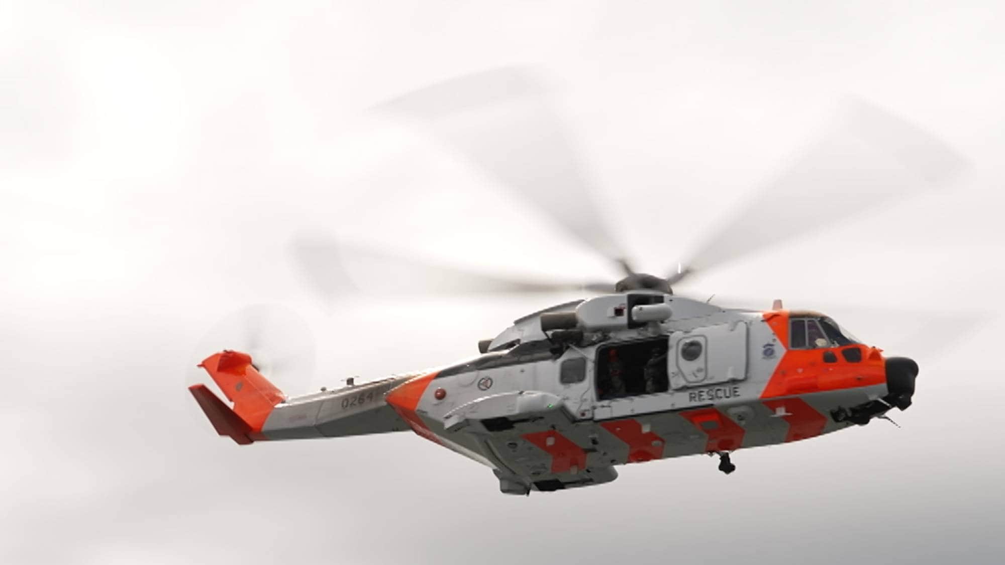 Et redningshelikopter er i bevegelse, svevende over et åpent landskap under en skyfull himmel. Helikopteret er malt i hvitt og oransje, med merket "RESCUE" synlig på siden. Rotorbladene er i aksjon, noe som gir bildets dynamikk. Omgivelsene inkluderer grønt landskap, men detaljene er uklare på grunn av avstand. (Bildebeskrivelsen er laget av en KI-tjeneste)