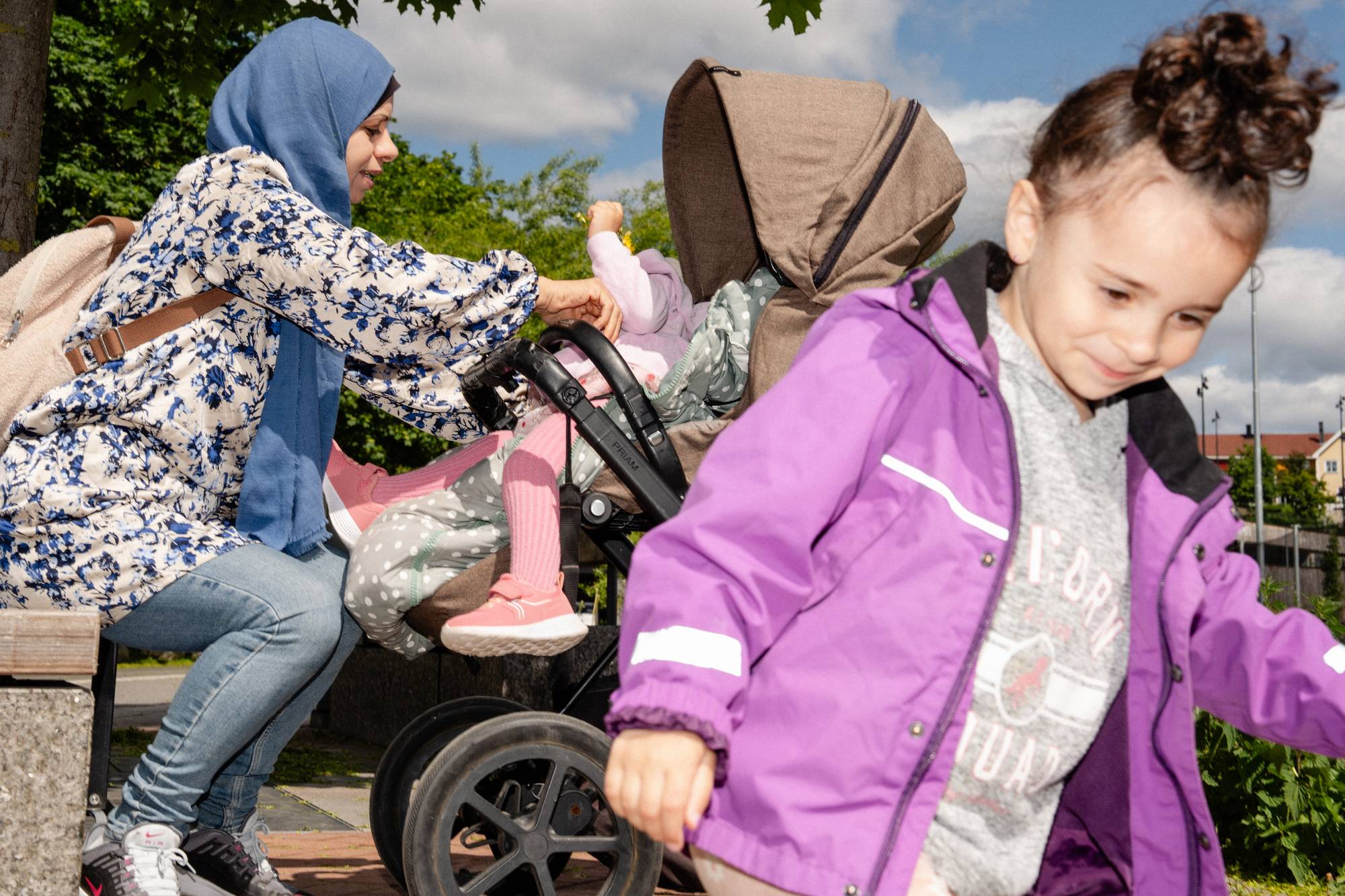 En kvinne med hijab ordner med et barn i en barnevogn. Et annet barn ses i forgrunnen, med et lite smil og lilla jakke.