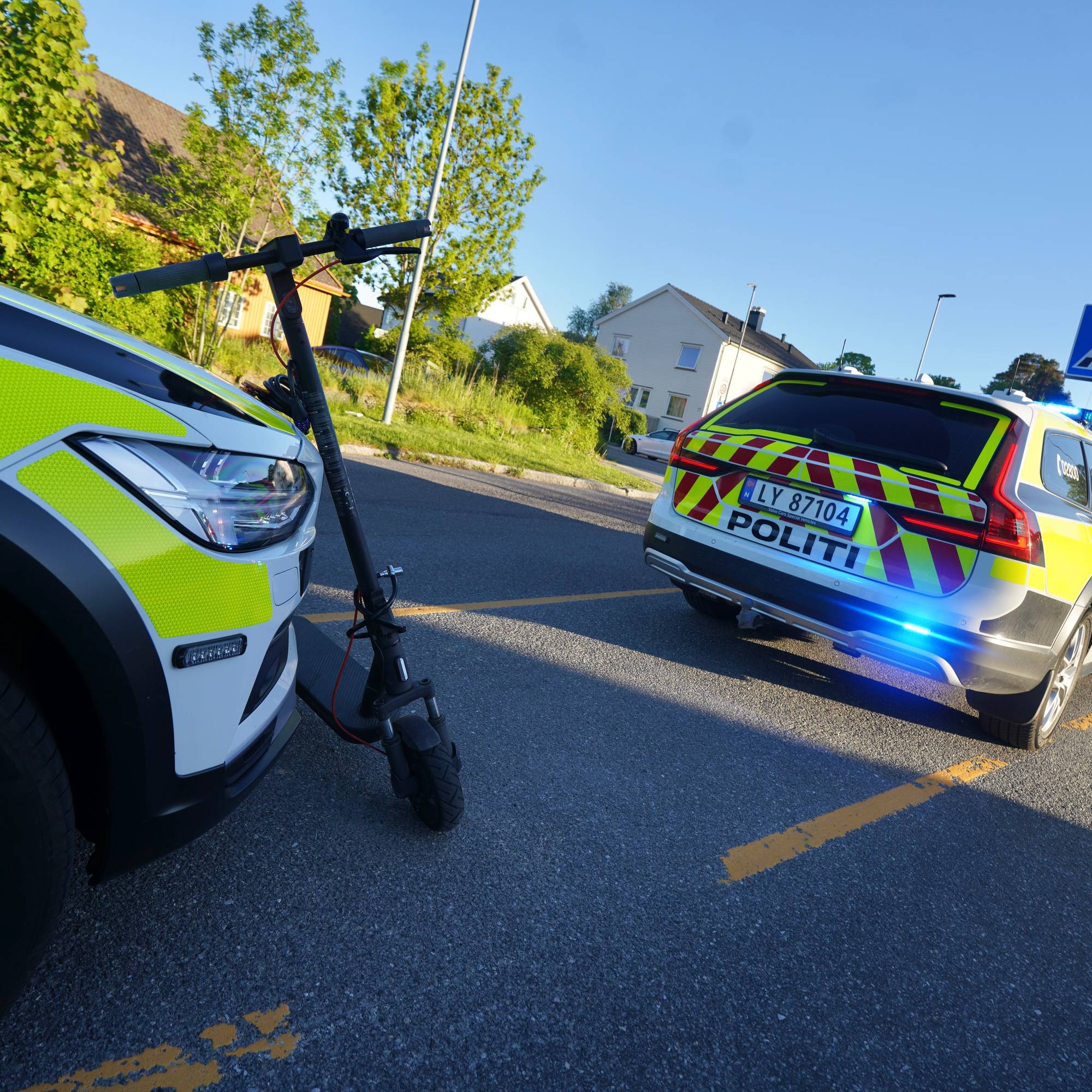 En elsparkesykkel står ved siden av en politibil på en gate. En annen politibil står lenger bak med blålys på. Det er en krysning med gangfelt og et blått trafikkskilt med gangikon. Omgivelsene består av bebyggelse og trær. (Bildebeskrivelsen er laget av en KI-tjeneste)