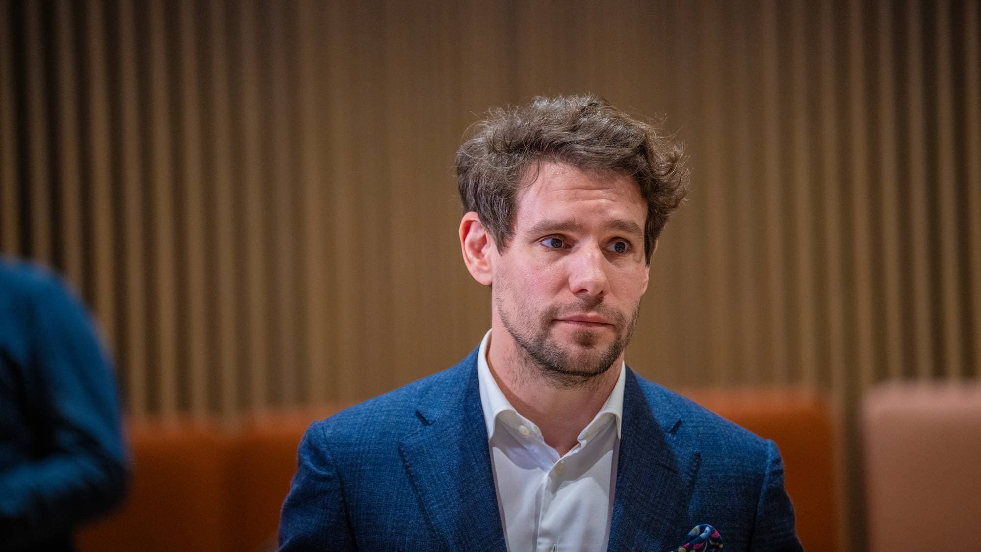 En mann i dress sitter foran en vegg av trepaneler. Han har brunt, krøllete hår og ser til siden. I bakgrunnen er det to personer til som sitter. (Bildebeskrivelsen er laget av en KI-tjeneste)