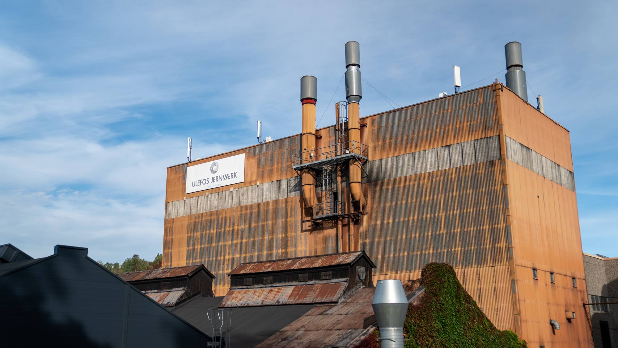 Fabrikkbygget til Ulefos Jernværk med høye piper og rustfarget fasade under en blå himmel. (Bildebeskrivelsen er laget av en KI-tjeneste)