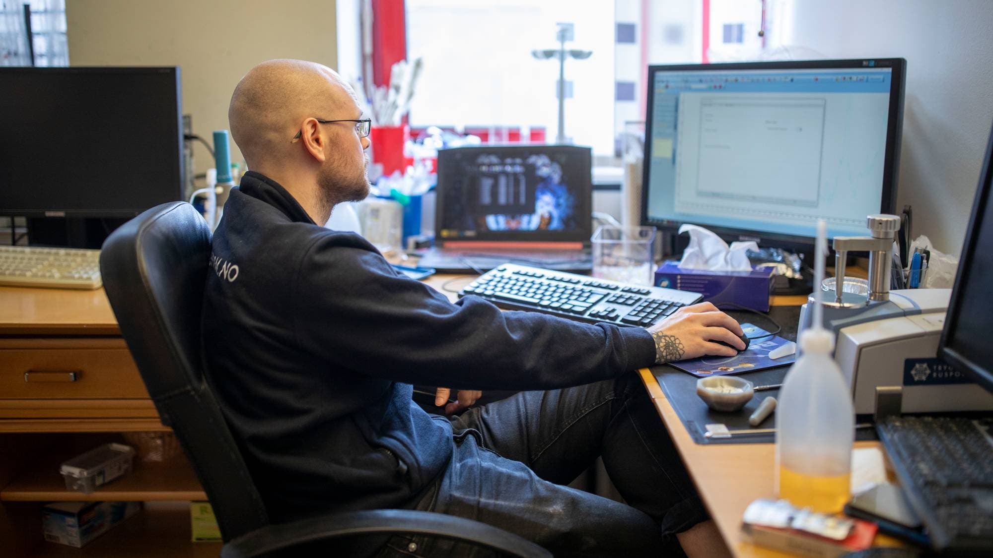 En mann med barbert hode og briller sitter ved et skrivebord og bruker en datamus. Mannen har på seg en mørk genser og jeans. Skrivebordet er utstyrt med flere dataskjermer, tastatur og annet utstyr. (Bildebeskrivelsen er laget av en KI-tjeneste)
