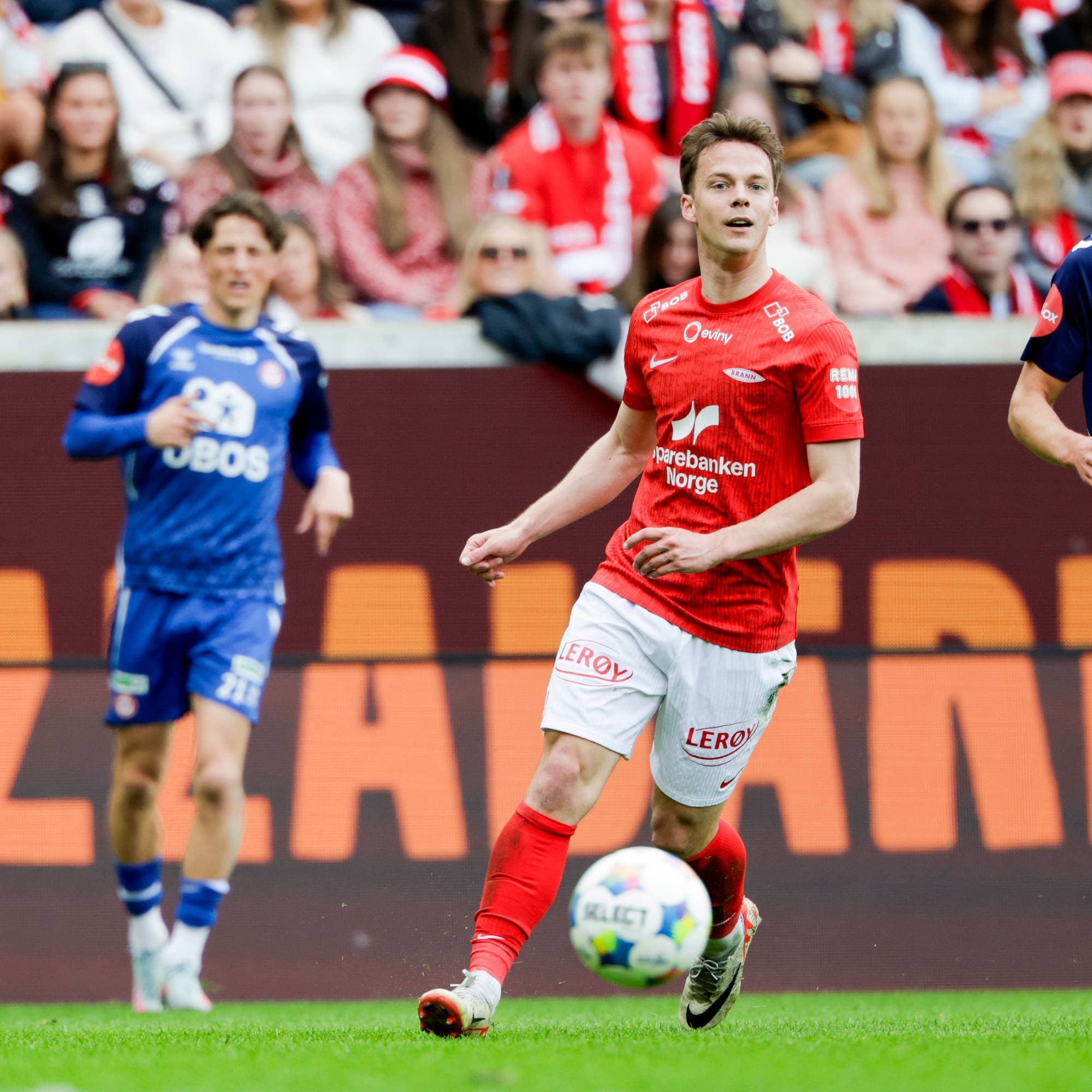 To fotballspillere er på en gressbane. Spilleren til venstre har på seg en blå drakt, og spilleren til høyre har på seg en rød drakt. Begge spillerne ser ut til å være i aksjon under en kamp. Bak spillerne sitter tilskuere på tribunen. (Bildebeskrivelsen er laget av en KI-tjeneste)