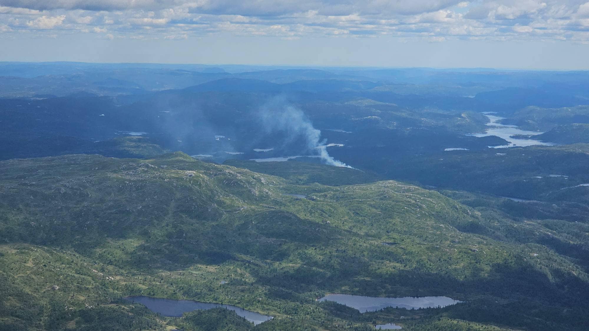 Røyk stiger opp fra et skogområde i et kupert fjellandskap sett fra luften. (Bildebeskrivelsen er laget av en KI-tjeneste)