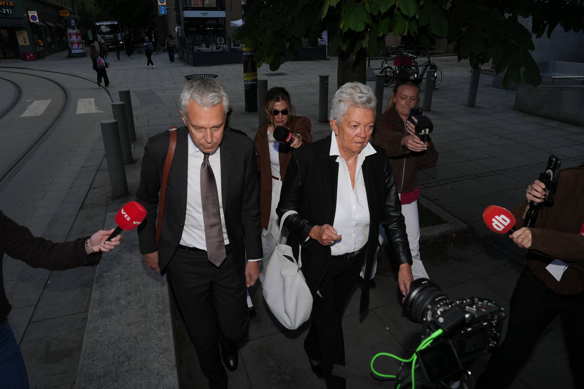 En mann i dress og en kvinne i dressjakke går utendørs, omringet av journalister.
Flere journalister med mikrofoner, kameraer og telefoner dokumenterer.
Det er sporvognsskinner og fortau i bakgrunnen.
En person i mørk brun jakke holder en mikrofon mot kvinnen. (Bildebeskrivelsen er laget av en KI-tjeneste)